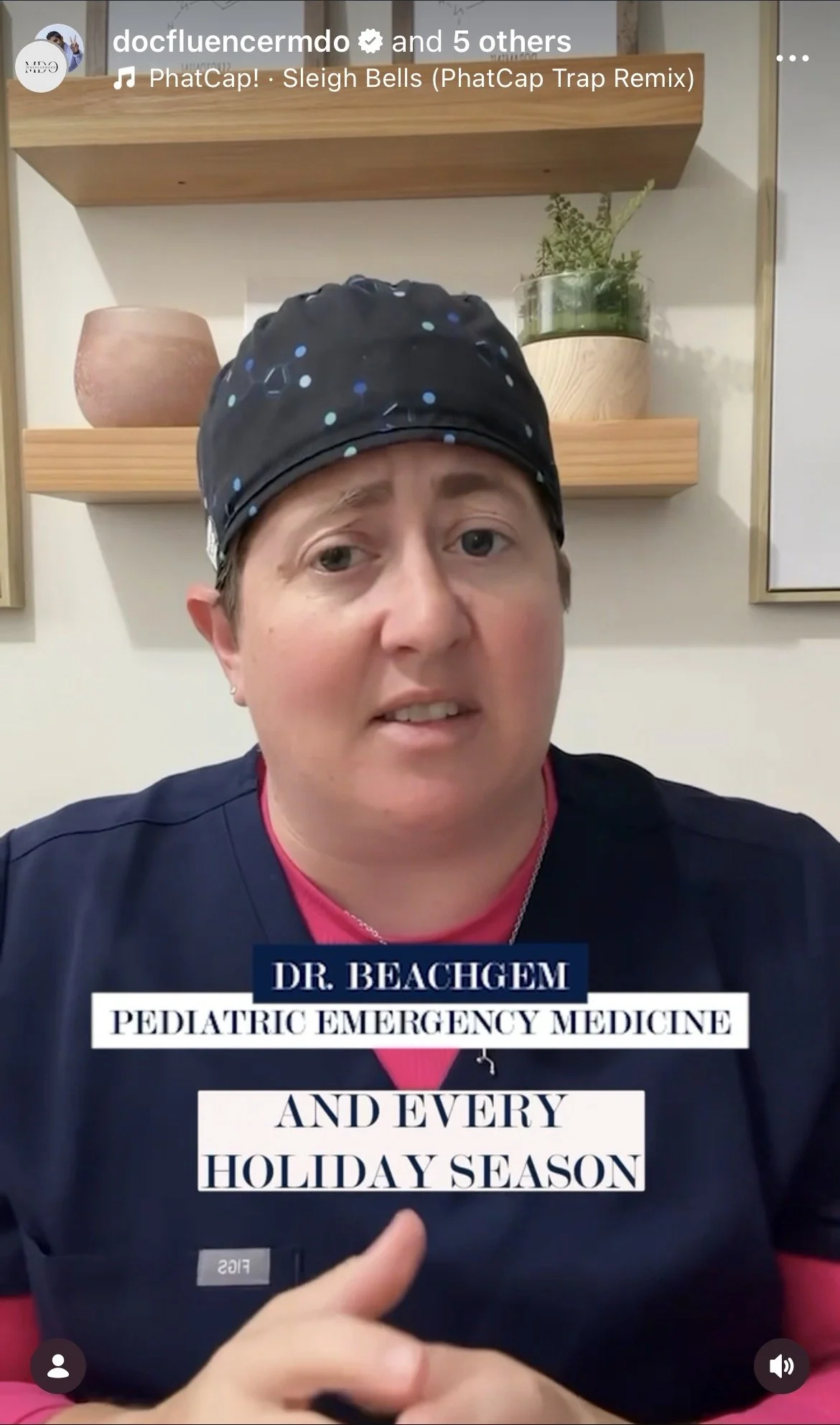 A woman wearing a black scrub top with a pink shirt underneath and a dark scrub cap with blue polka dots. She is speaking and appears to be a pediatric emergency medicine doctor. Behind her are wooden shelves with a pink vase, a green plant, and a framed picture.