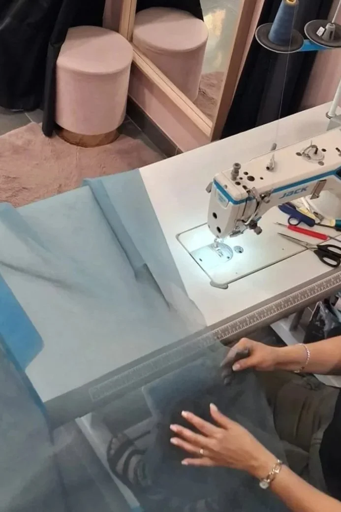 Tavolo da lavoro con macchina da cucire, forbici, e tessuto blu, in un ambiente domestico o laboratorio di sartoria.