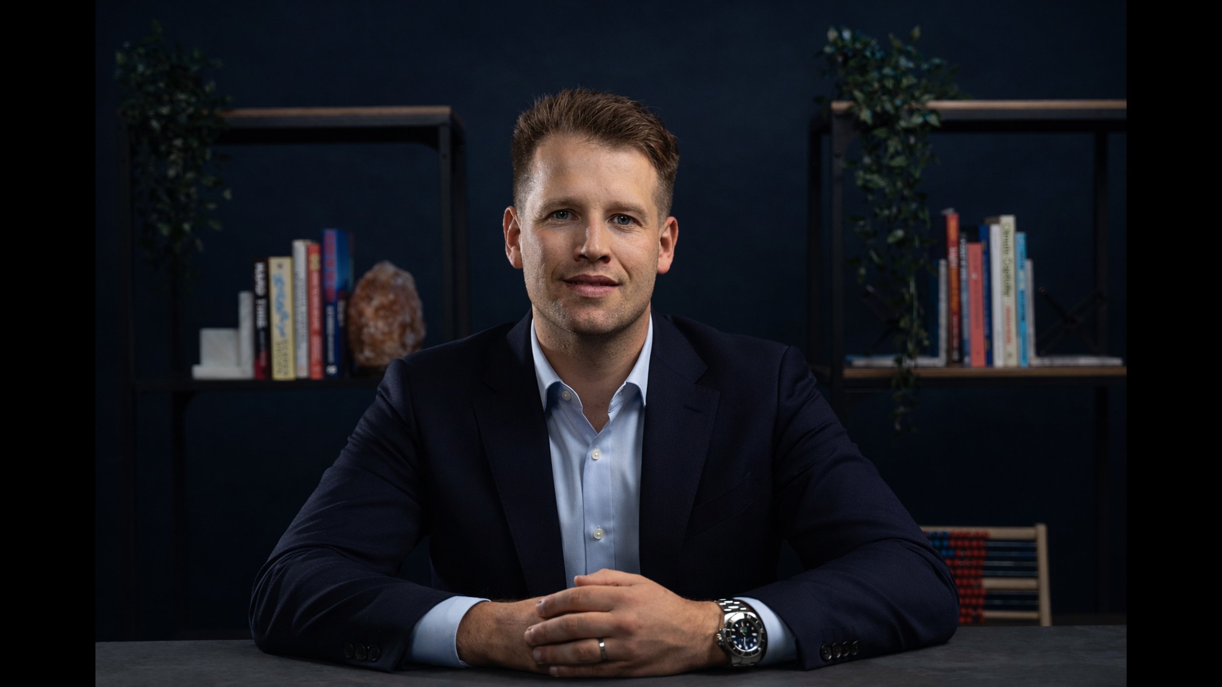 A man in a dark suit and white shirt sitting at a table with hands folded, in a dimly lit room with dark blue walls, bookshelf behind him with colorful books and decorative items.