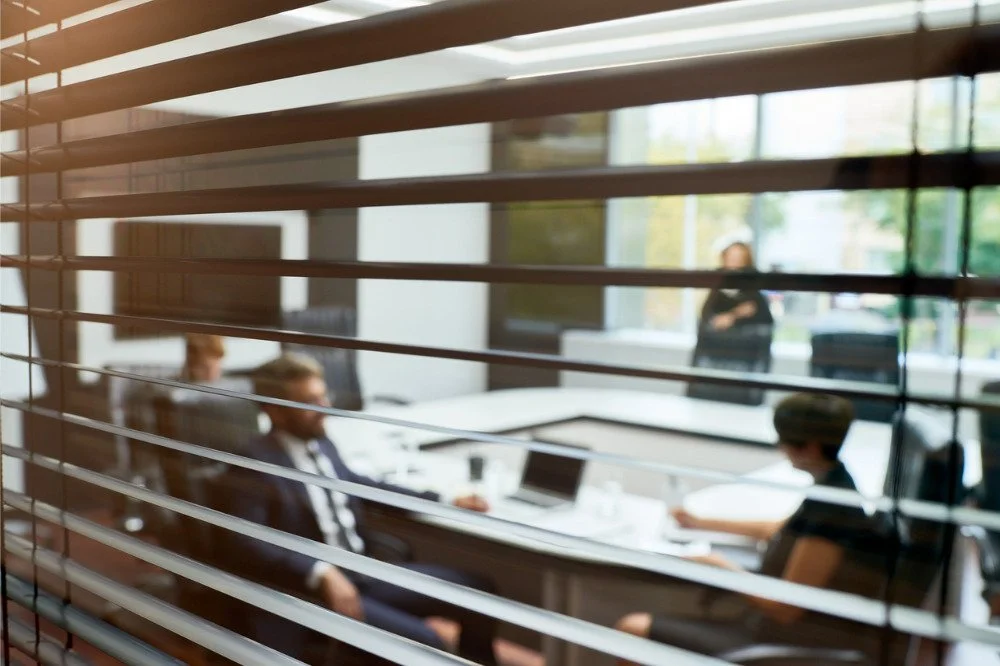 Business meeting room viewed through window blinds, with people working and discussing at a table.