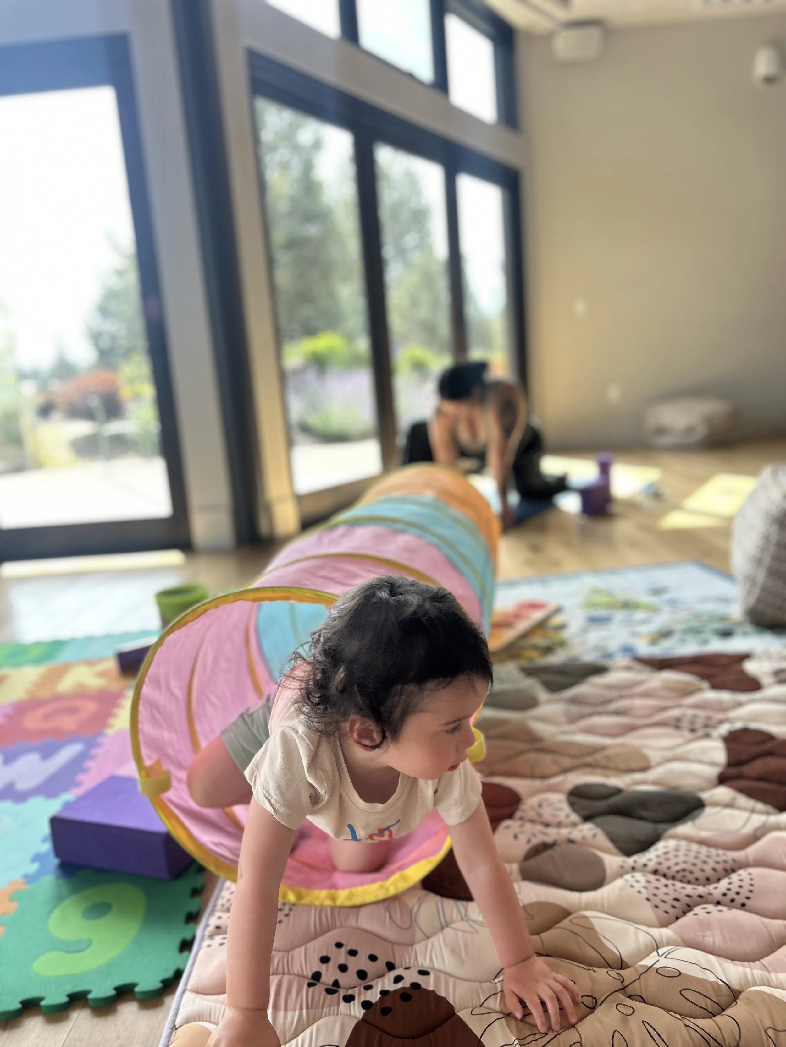 Child crawling through a colorful play tunnel on a soft padded mat in a bright room with large windows and a woman in the background.