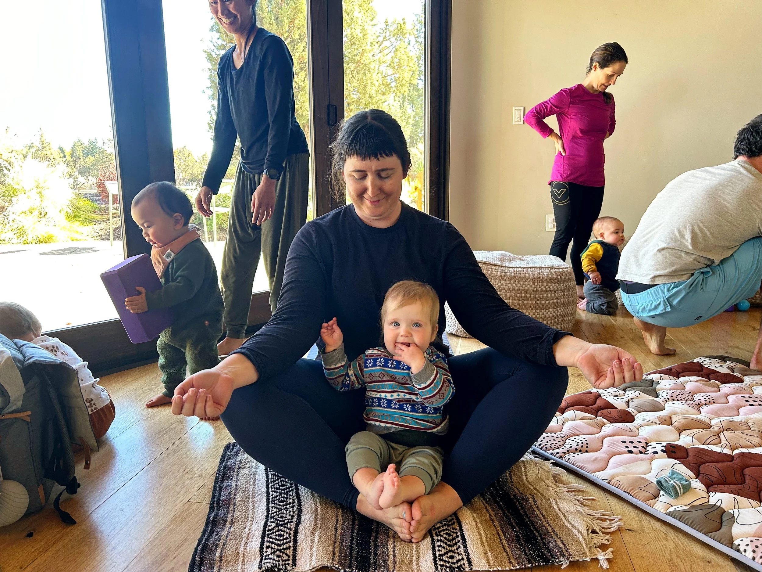 A group of women and children in a sunlit room, with one woman sitting cross-legged on a rug, meditating with a smiling baby on her lap, children playing and adults engaging in activities around her, large glass sliding doors revealing a sunny outdoor scene.