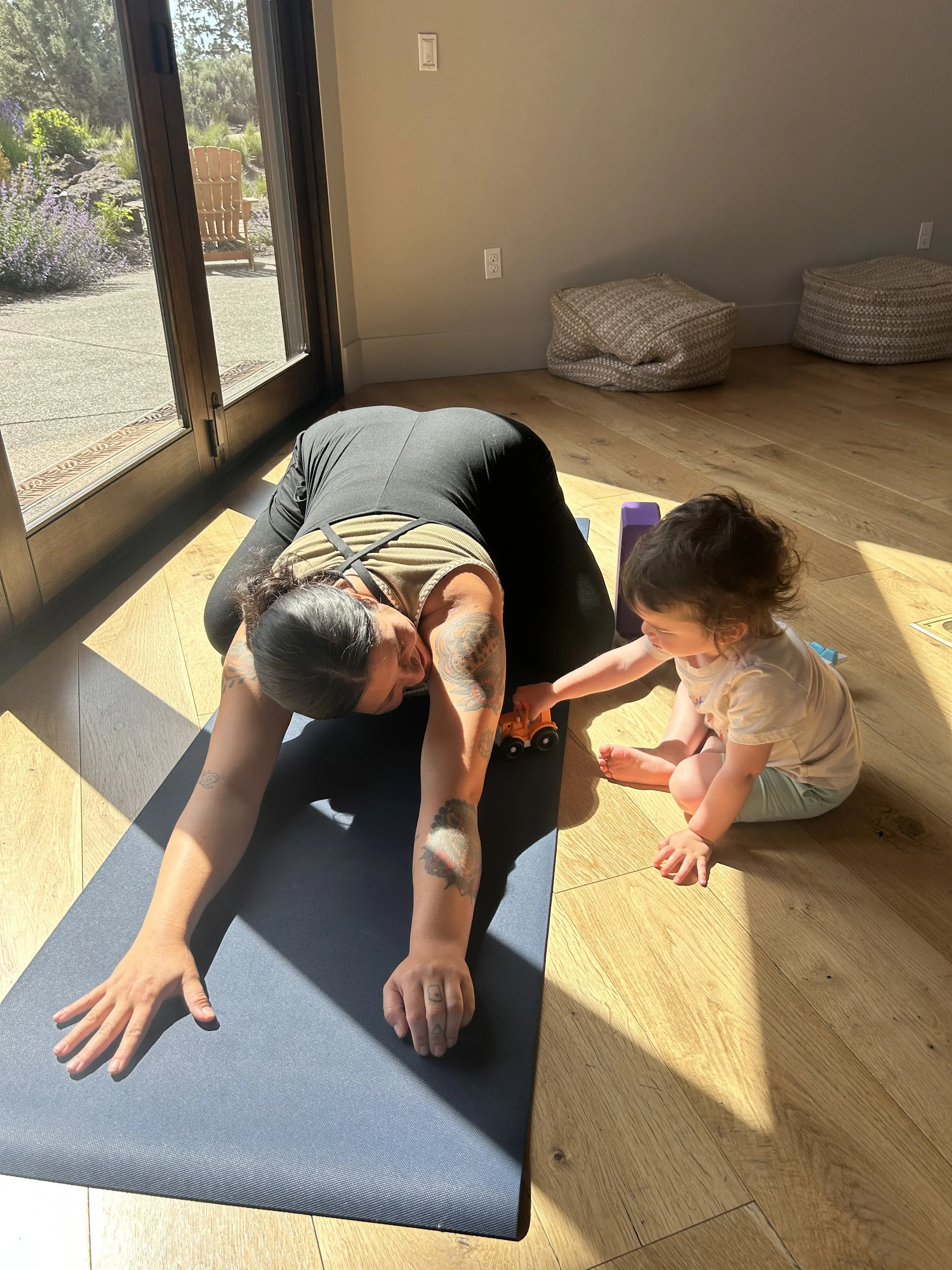 A woman practicing yoga on a mat inside a room with sunlight streaming in, while a young girl plays with a toy truck nearby.