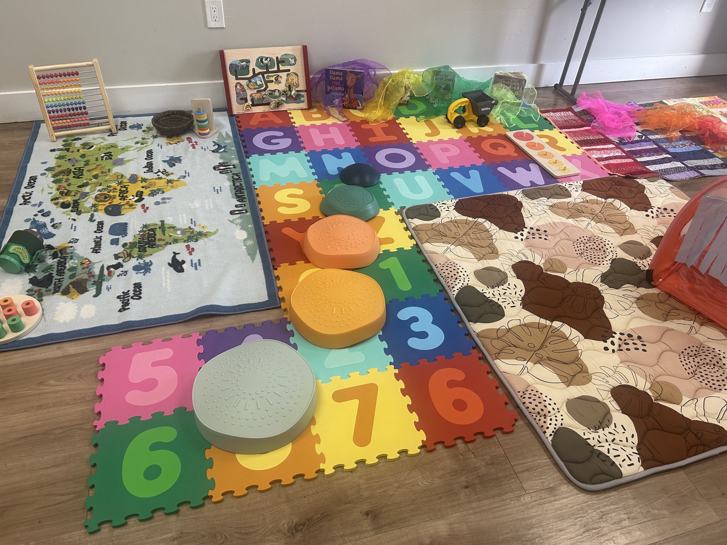 Colorful foam mats, plush animal pillows, and toys arranged in play area, including a small rug with jungle animals and word art, an abacus, and various textured stepping stones.