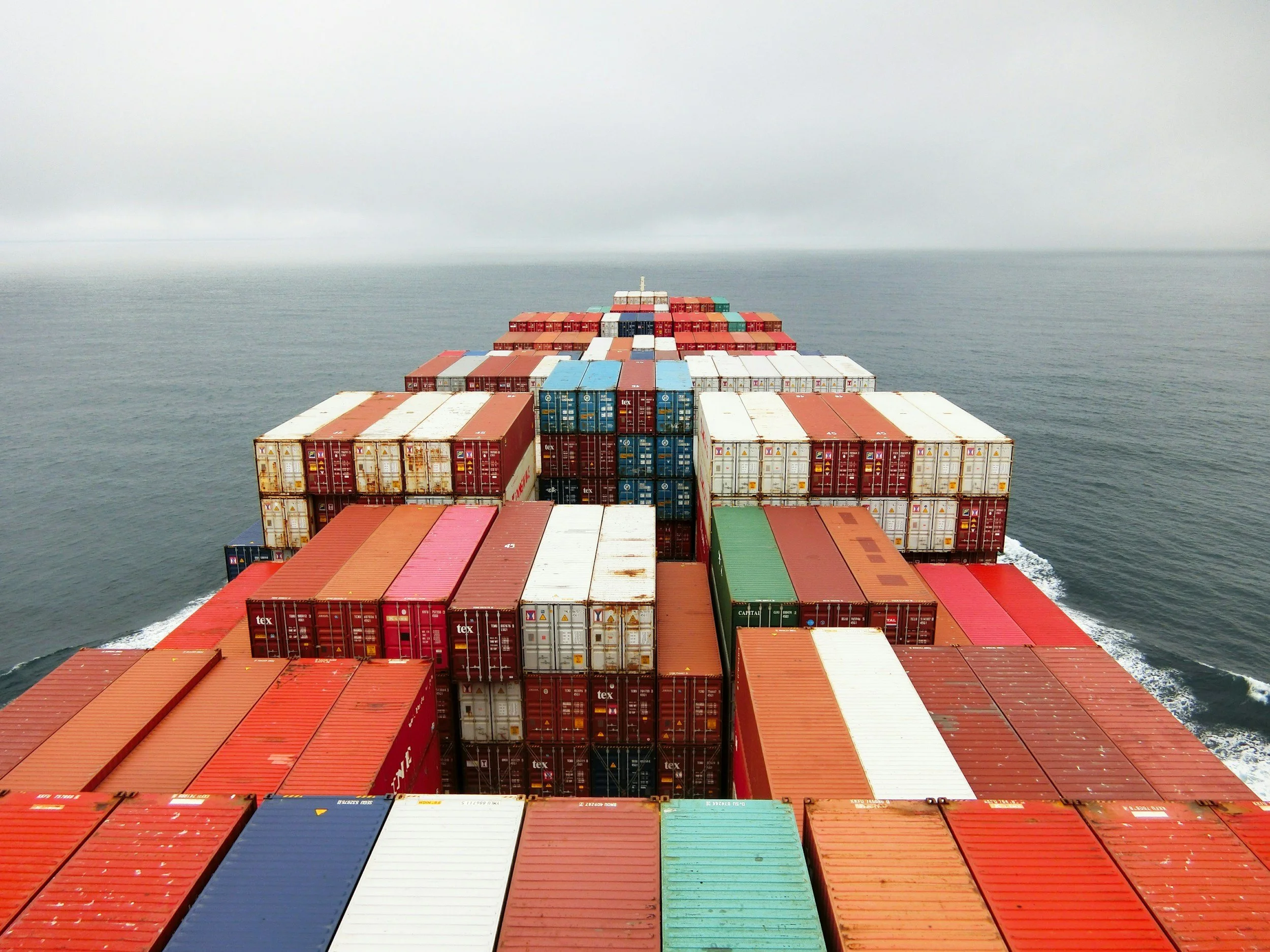 View from a cargo ship with stacked shipping containers in various colors sailing in the ocean under a cloudy sky.