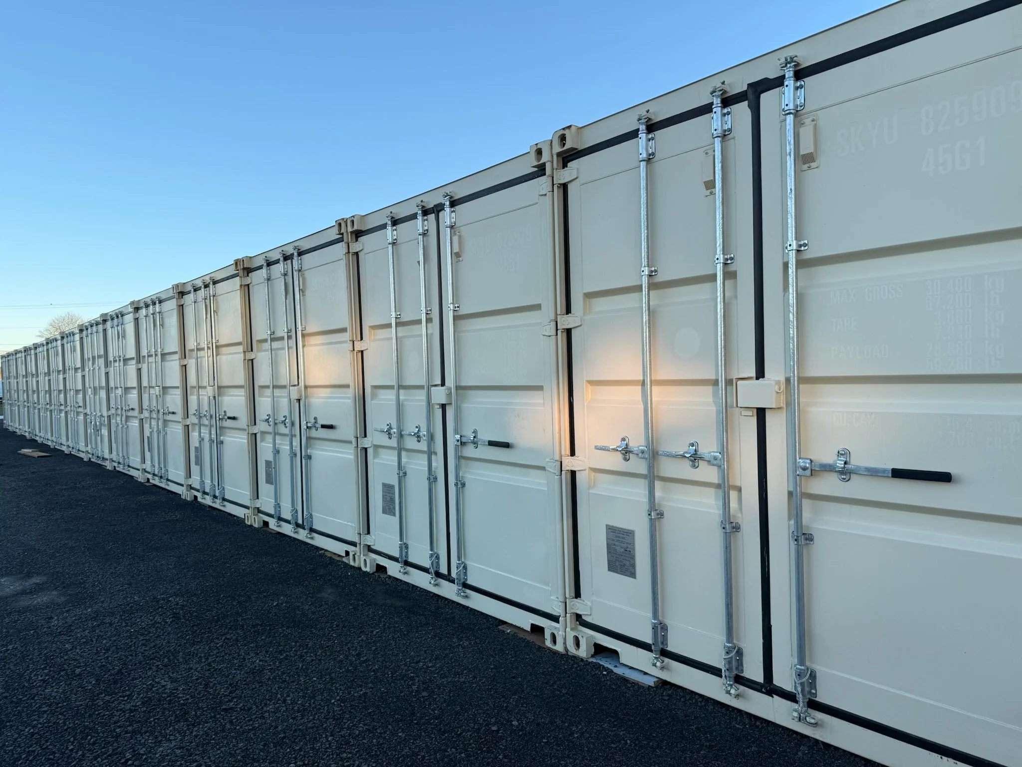 A long row of beige shipping containers on black asphalt under a clear blue sky.