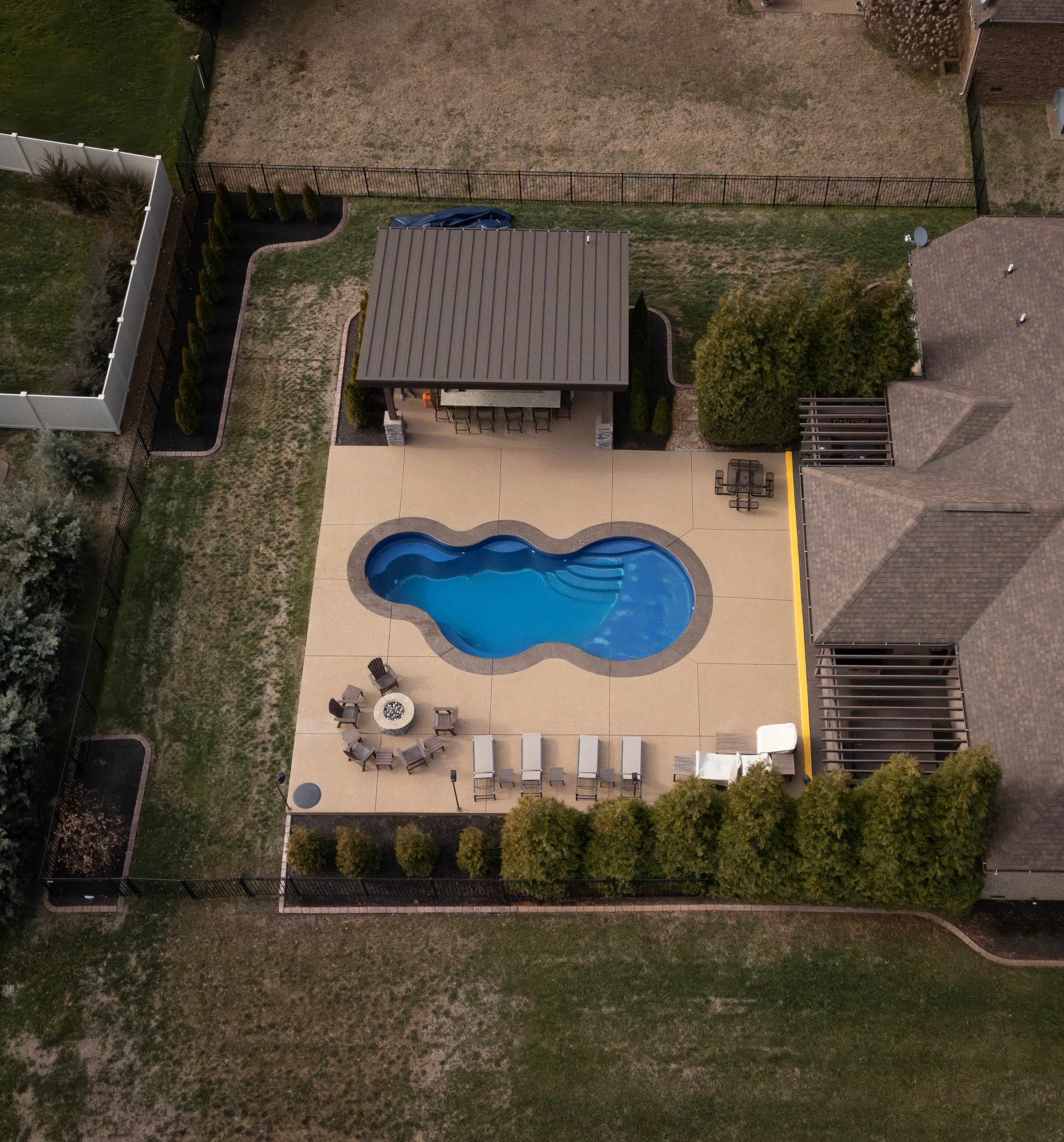 A backyard with a swimming pool, patio furniture, a small building with a bar, surrounded by a fence and neighboring houses.