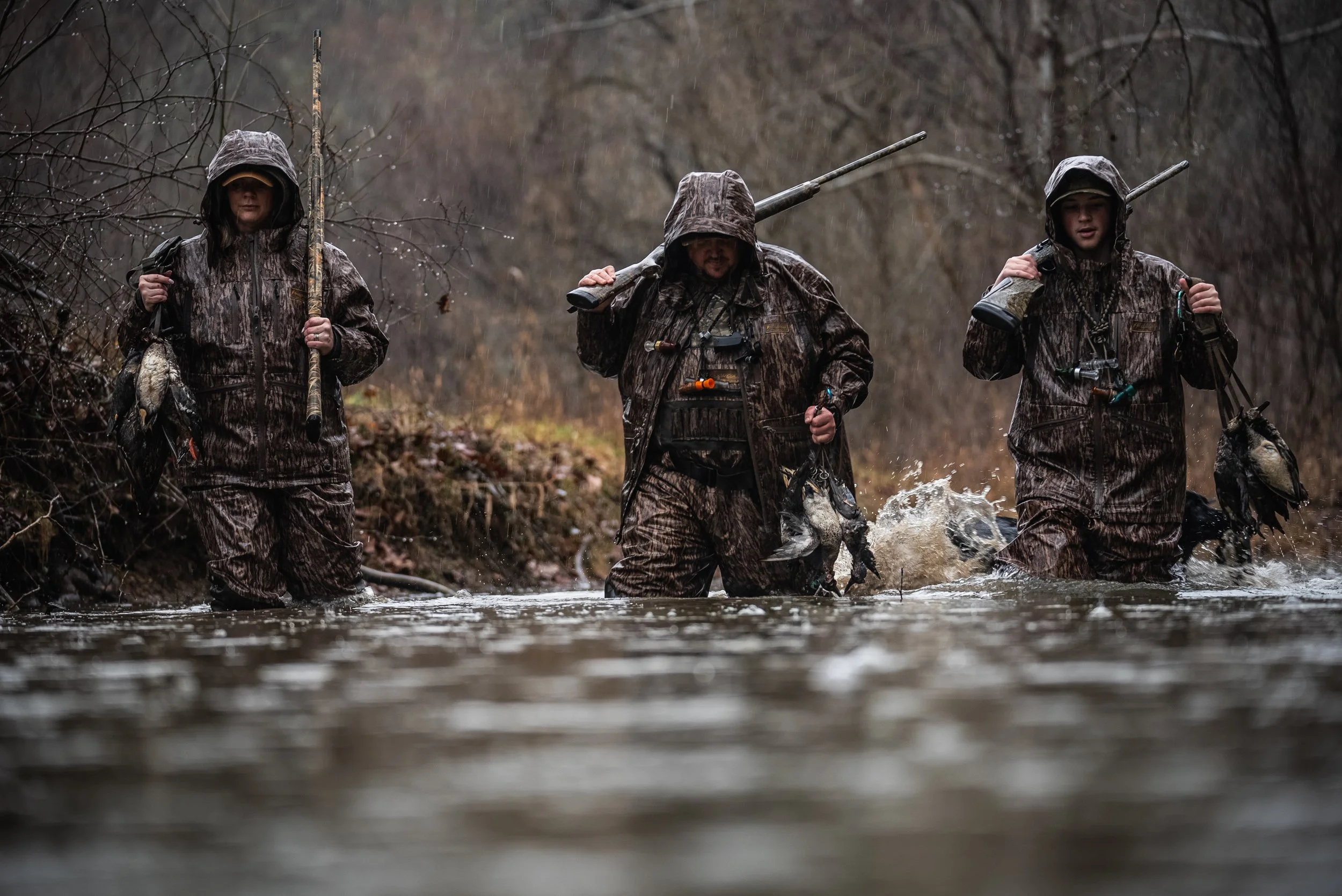 Three people in camouflage waterproof clothing wading through a river with fishing rods, carrying caught fish after a fishing trip.