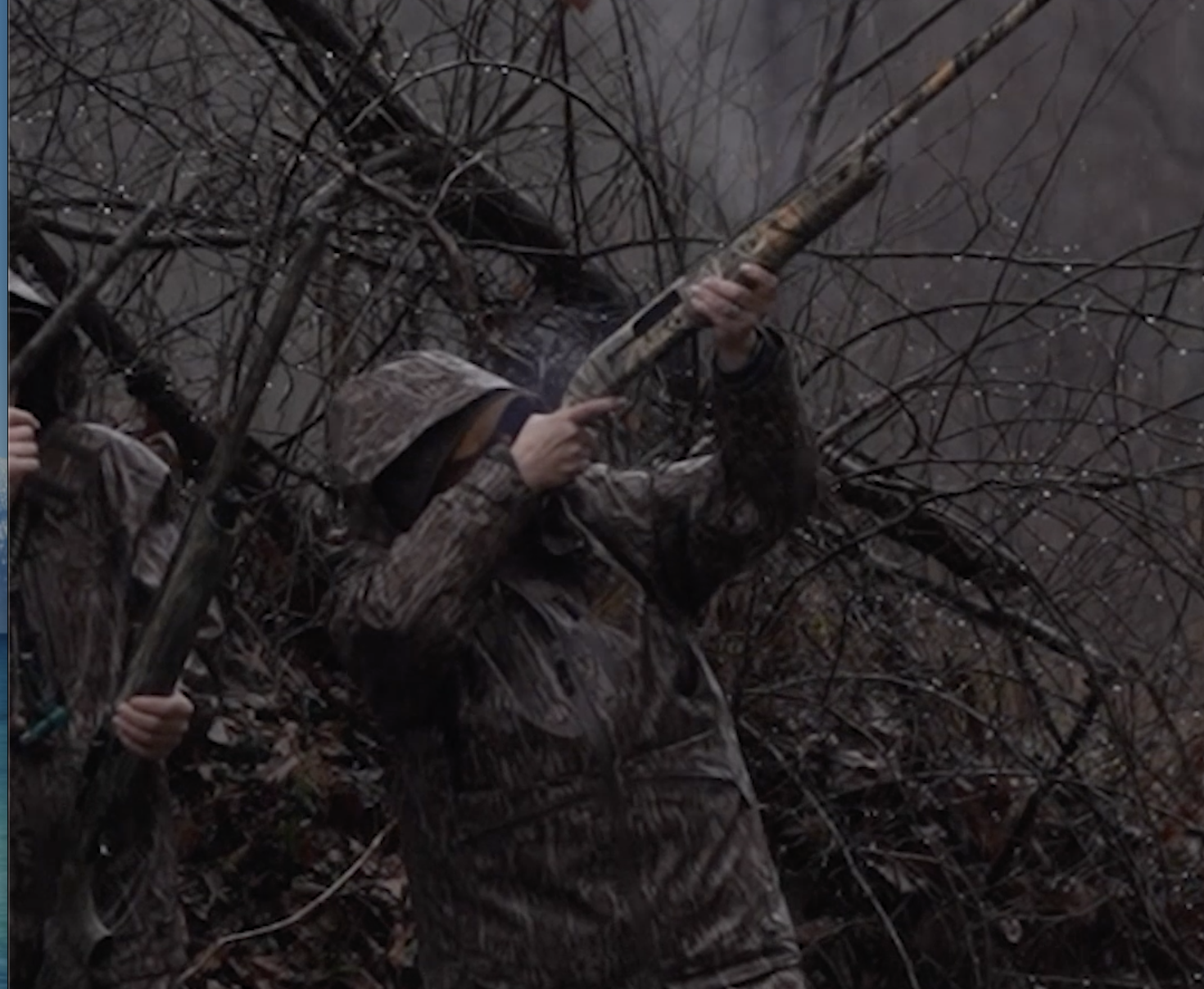 A person in camouflage clothing holding a shotgun among leafless branches near a body of water.