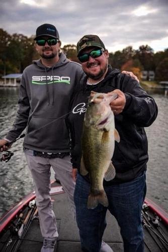 Two men on a boat outdoors, one holding a large fish, with trees and water in the background.