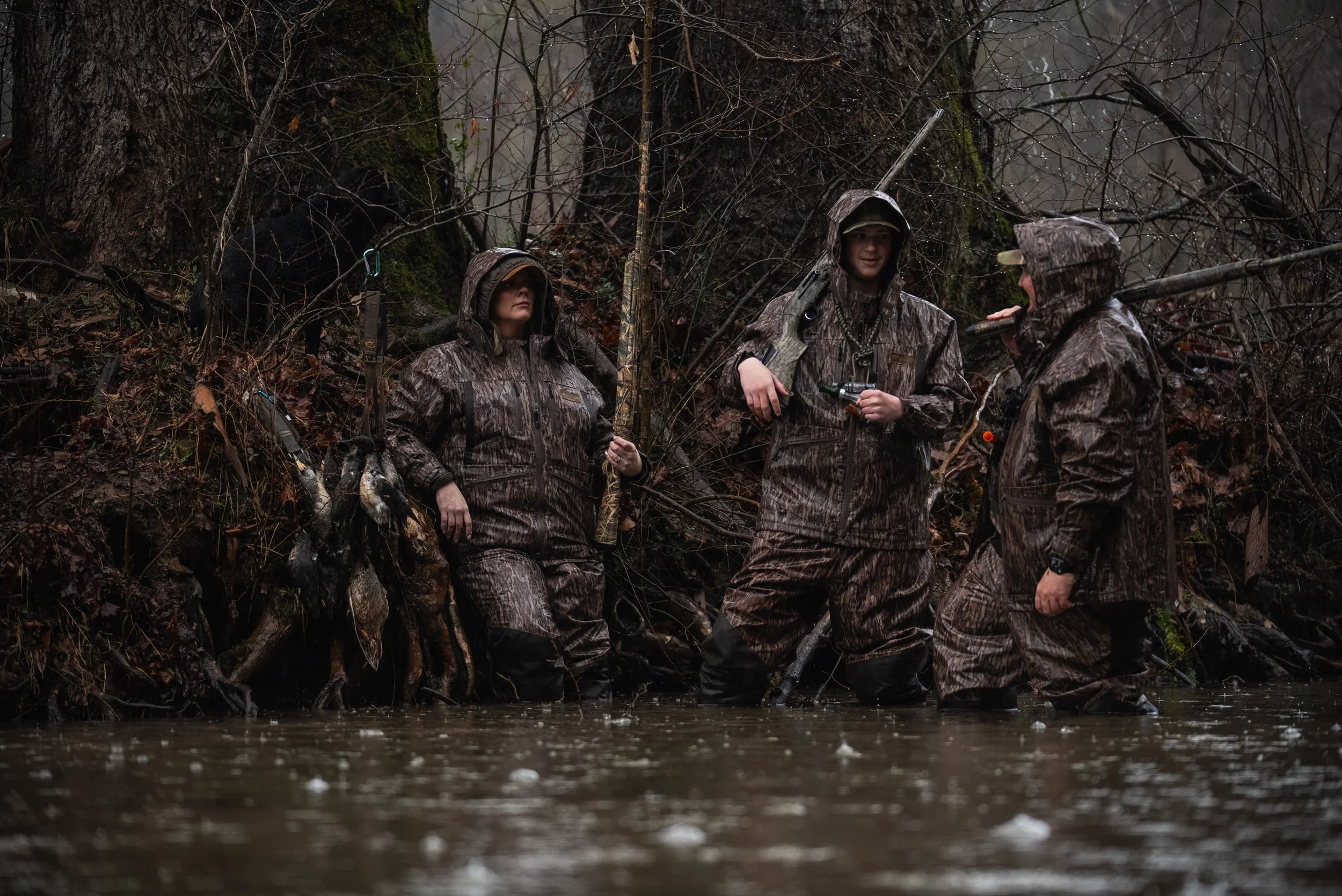 Four people dressed in camouflage waders and jackets standing in a shallow river, surrounded by leafless trees and branches.