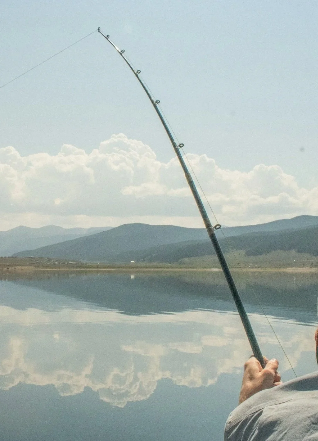 Person holding a fishing rod on a calm lake with mountains and cloudy sky in the background.