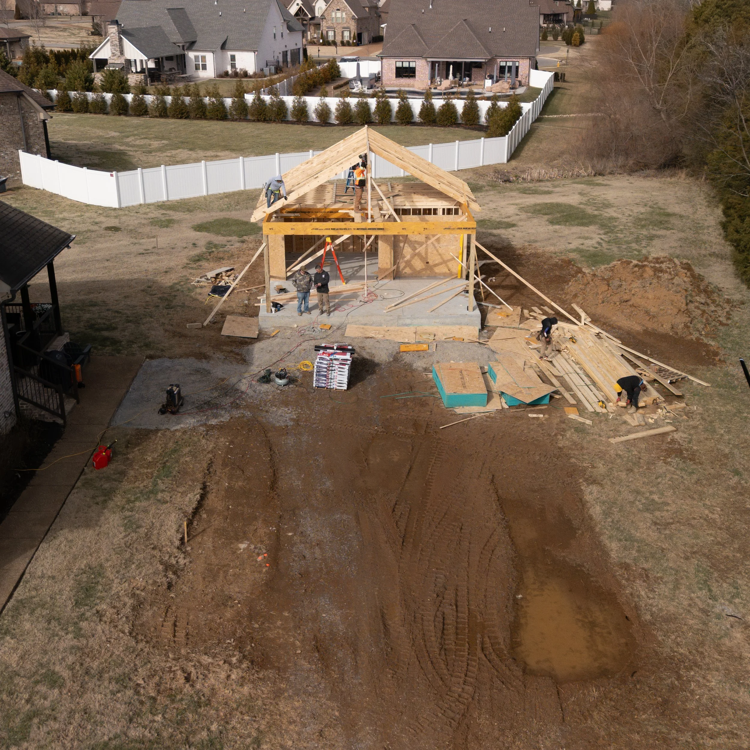 An aerial view of a house under construction with workers building the wooden framework. The construction site has scattered materials and equipment, with a muddy area and tire tracks in the foreground. Residential houses with fenced yards are visibl