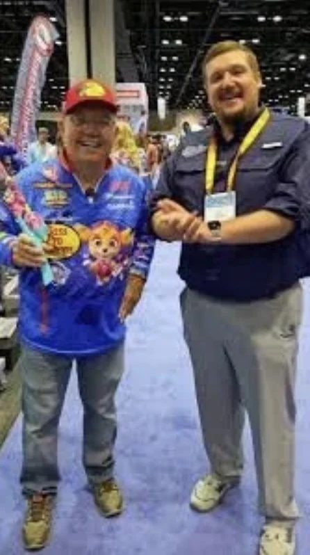 Two men shaking hands at a convention or trade show. One is wearing a colorful shirt with cartoon characters and a red cap, the other is wearing a navy shirt with a lanyard. The background shows other attendees and display booths.