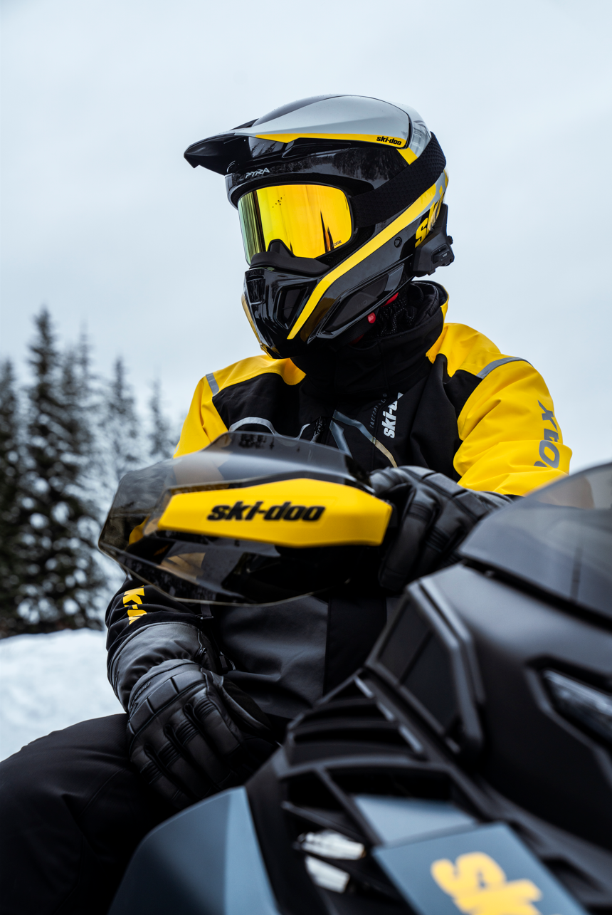 Person i gul og svart snowmobiltøy og hjelm med refleksjon, sitende på en svart og gul snowmobile i snødekt landskap med trær i bakgrunnen.