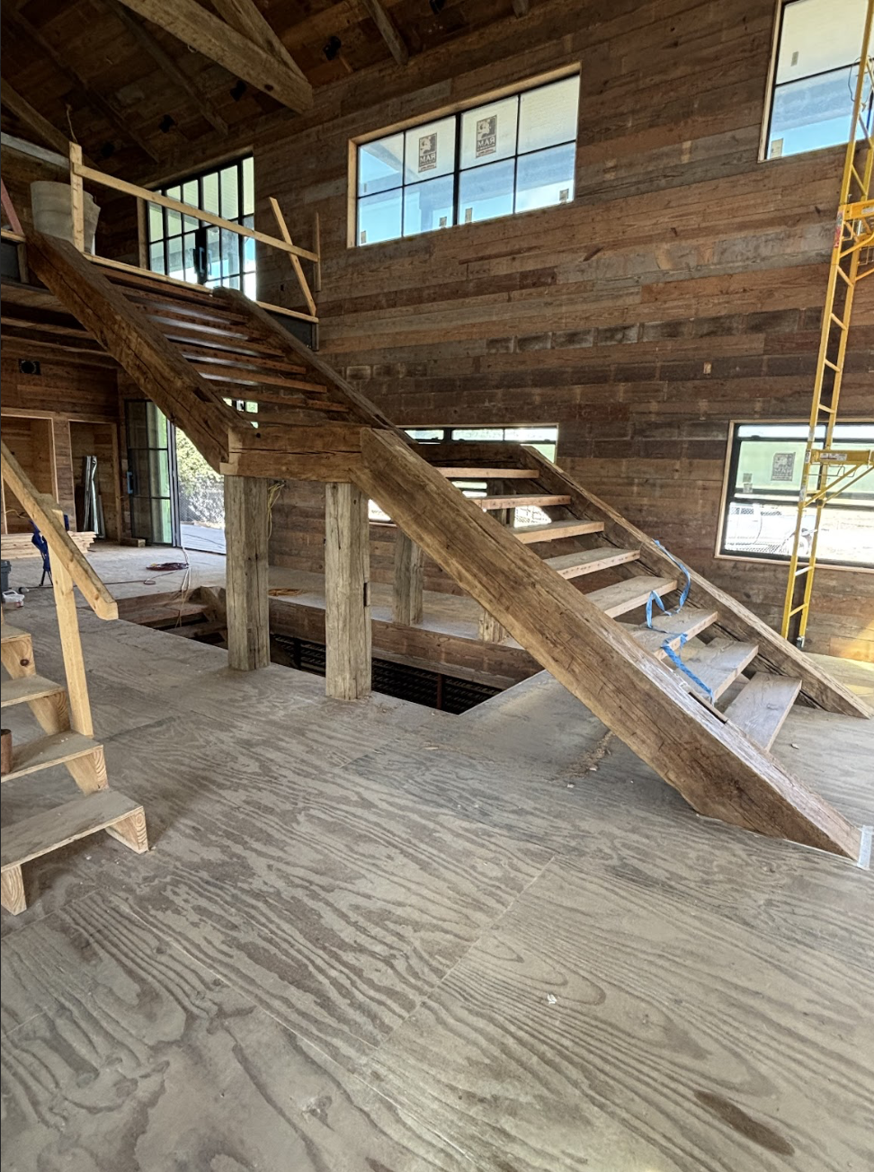 Interior view of a wooden staircase under construction in a rustic-style room with wood-paneled walls and large windows.