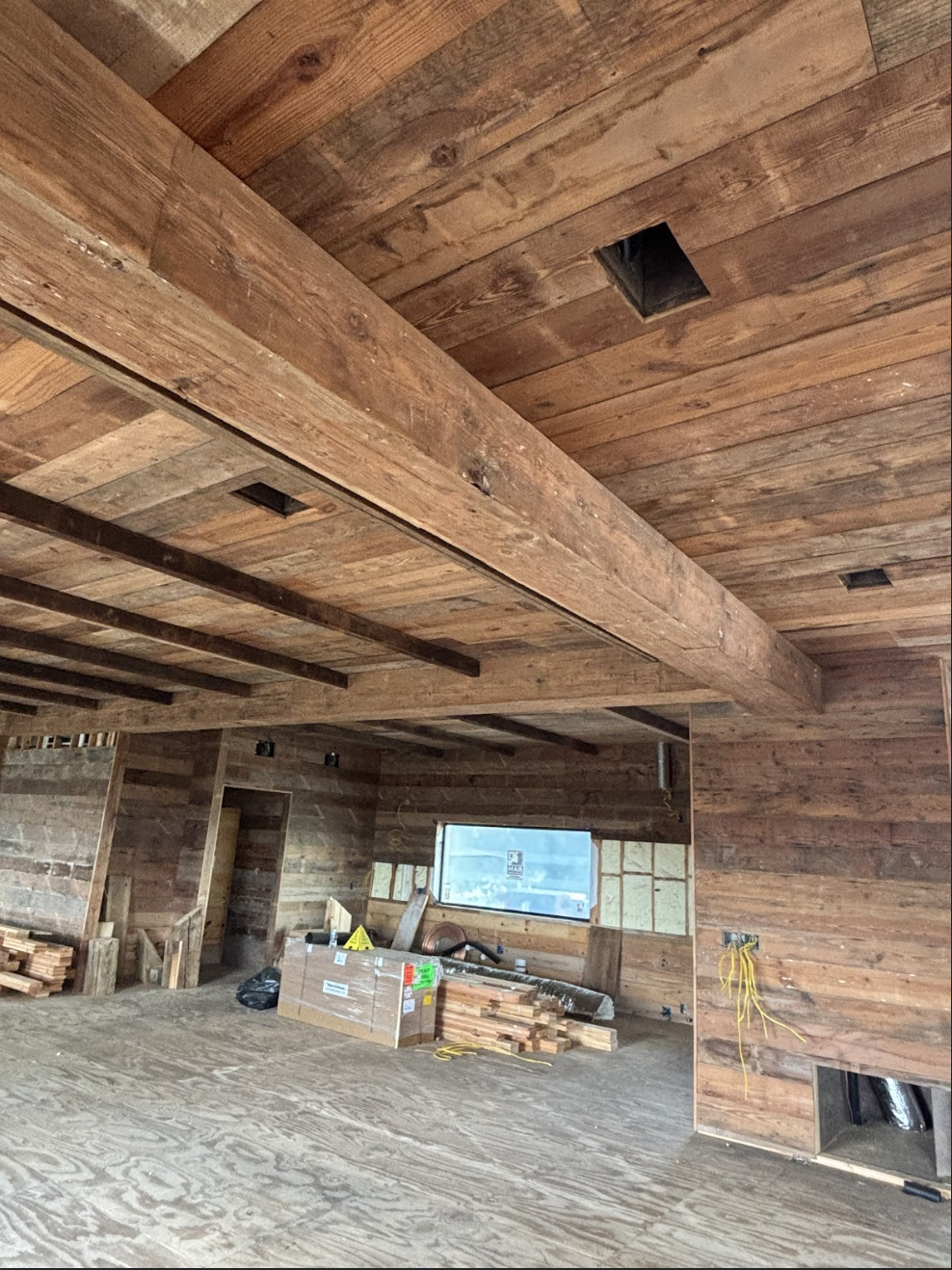 Interior of a building under construction with wooden walls and ceiling, exposed beams, a window, and construction materials and tools scattered on the floor.