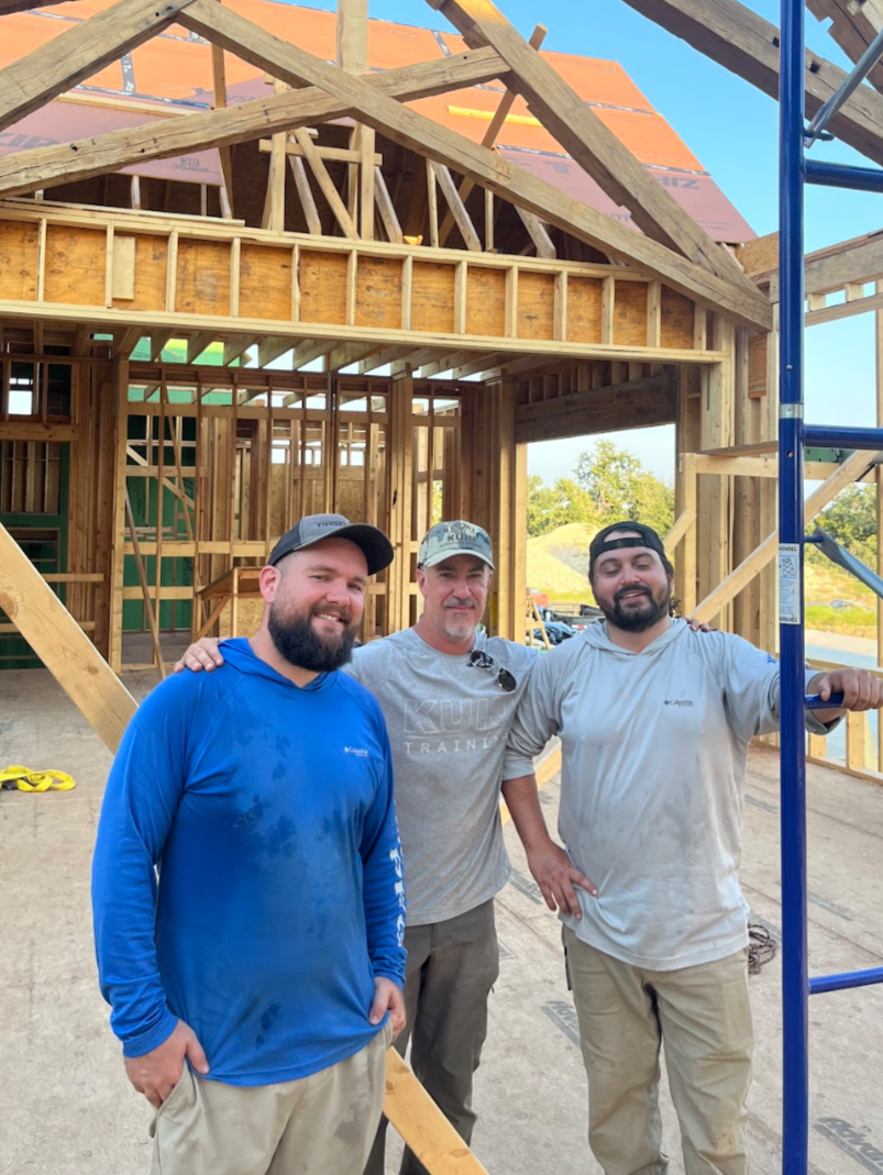 Three men smiling at the camera at a construction site of a wooden house or building with framed walls and roof structure under construction.