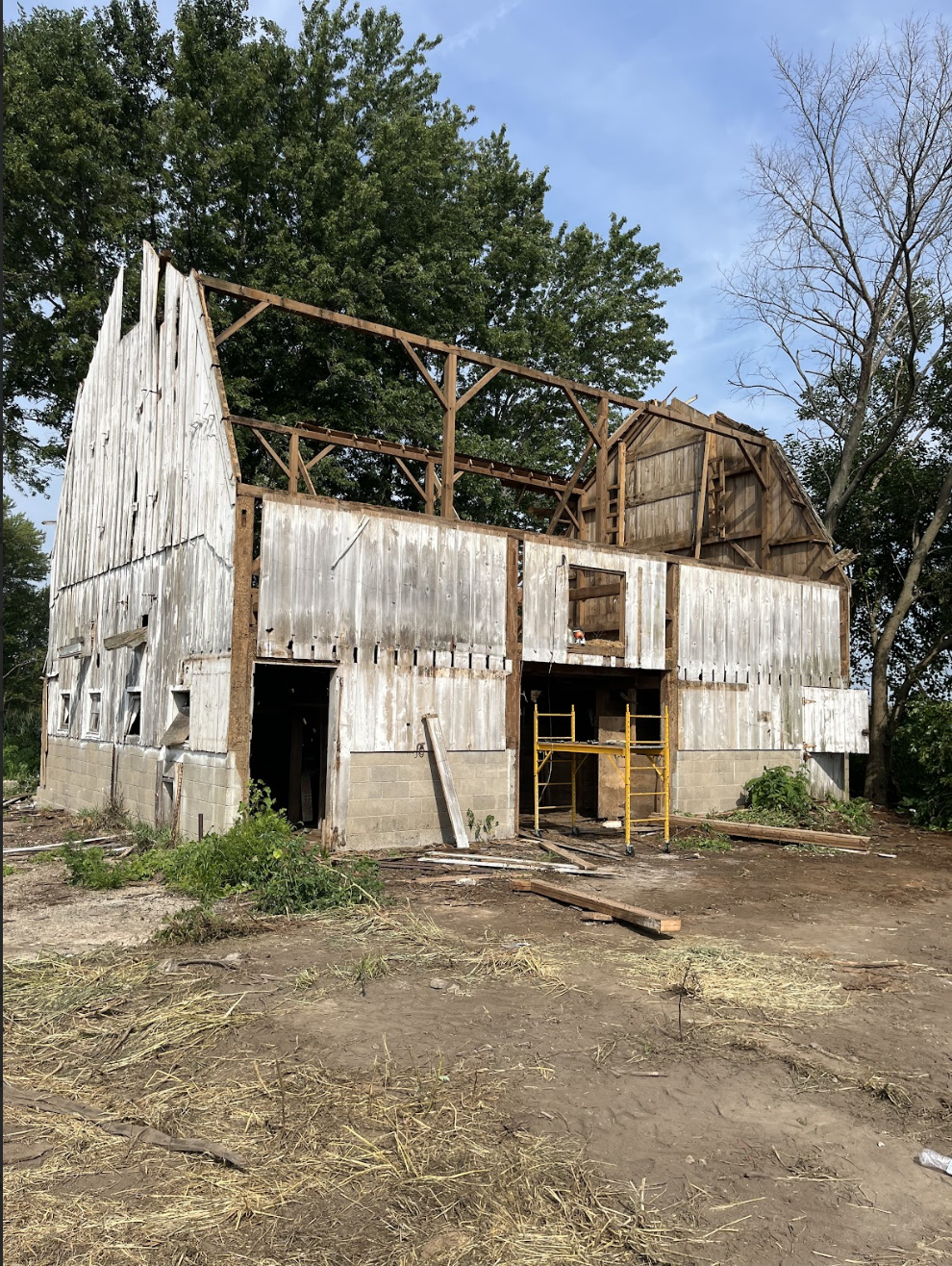 A partially constructed barn with a wooden frame and weathered siding, surrounded by trees and construction tools, on a dirt lot.