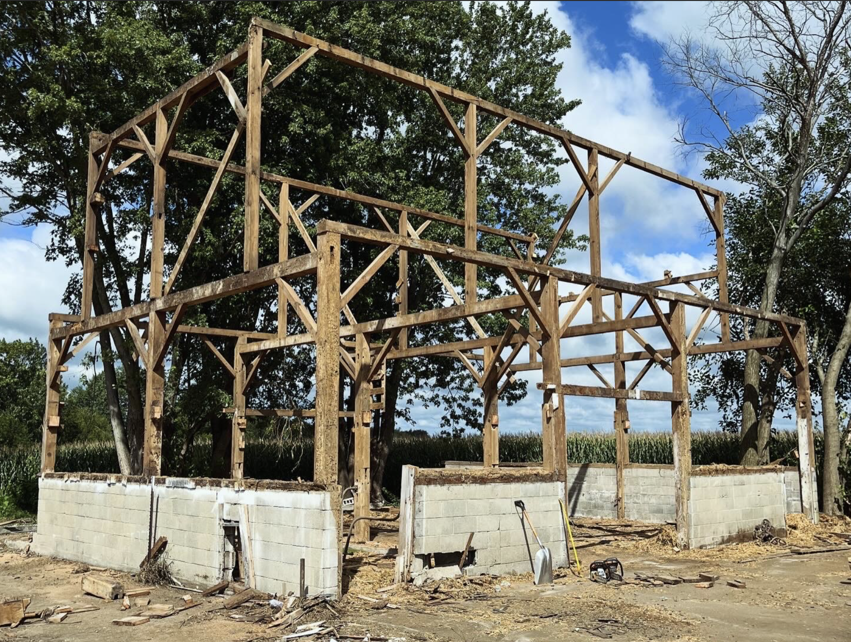 Wooden framework of a building under construction with cinder block walls at the base, surrounded by construction tools and equipment, with trees and a blue sky with clouds in the background.