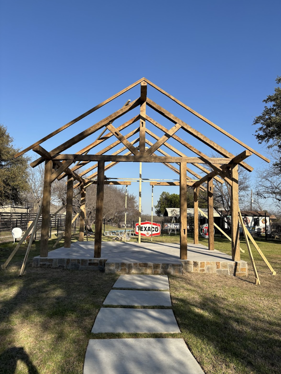 Wooden house frame under construction with a concrete walkway leading to it, set in a grassy area with trees and vintage signs in the background.