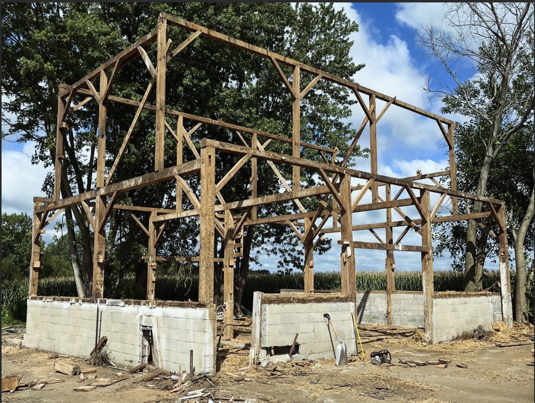 Wooden framing of a building under construction with concrete block walls, surrounded by trees and construction tools, under a partly cloudy sky.