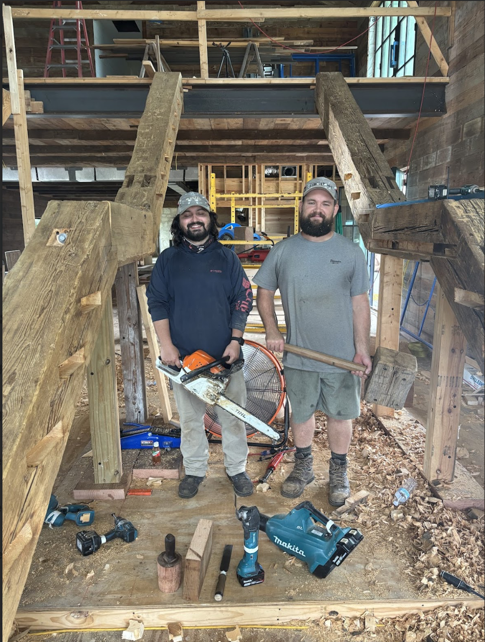 Two men smiling in a construction site, surrounded by tools and wooden structures, with unfinished woodwork and progress in the background.