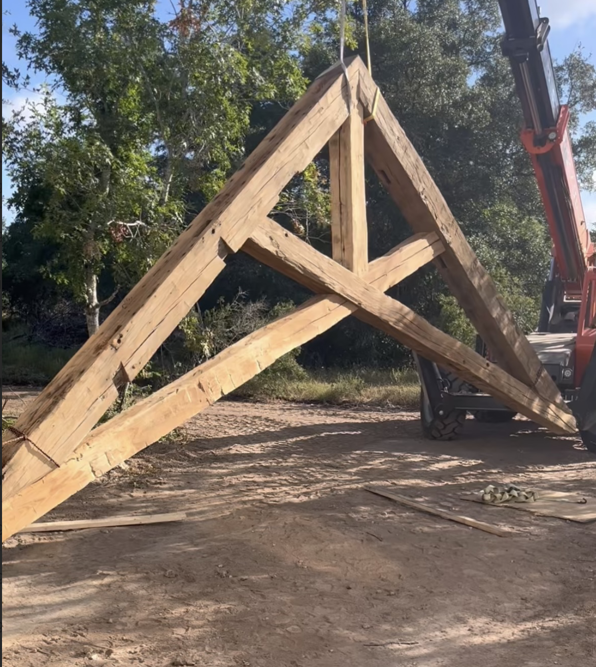 A large wooden triangular structure being hoisted by a crane on a dirt path with trees in the background.
