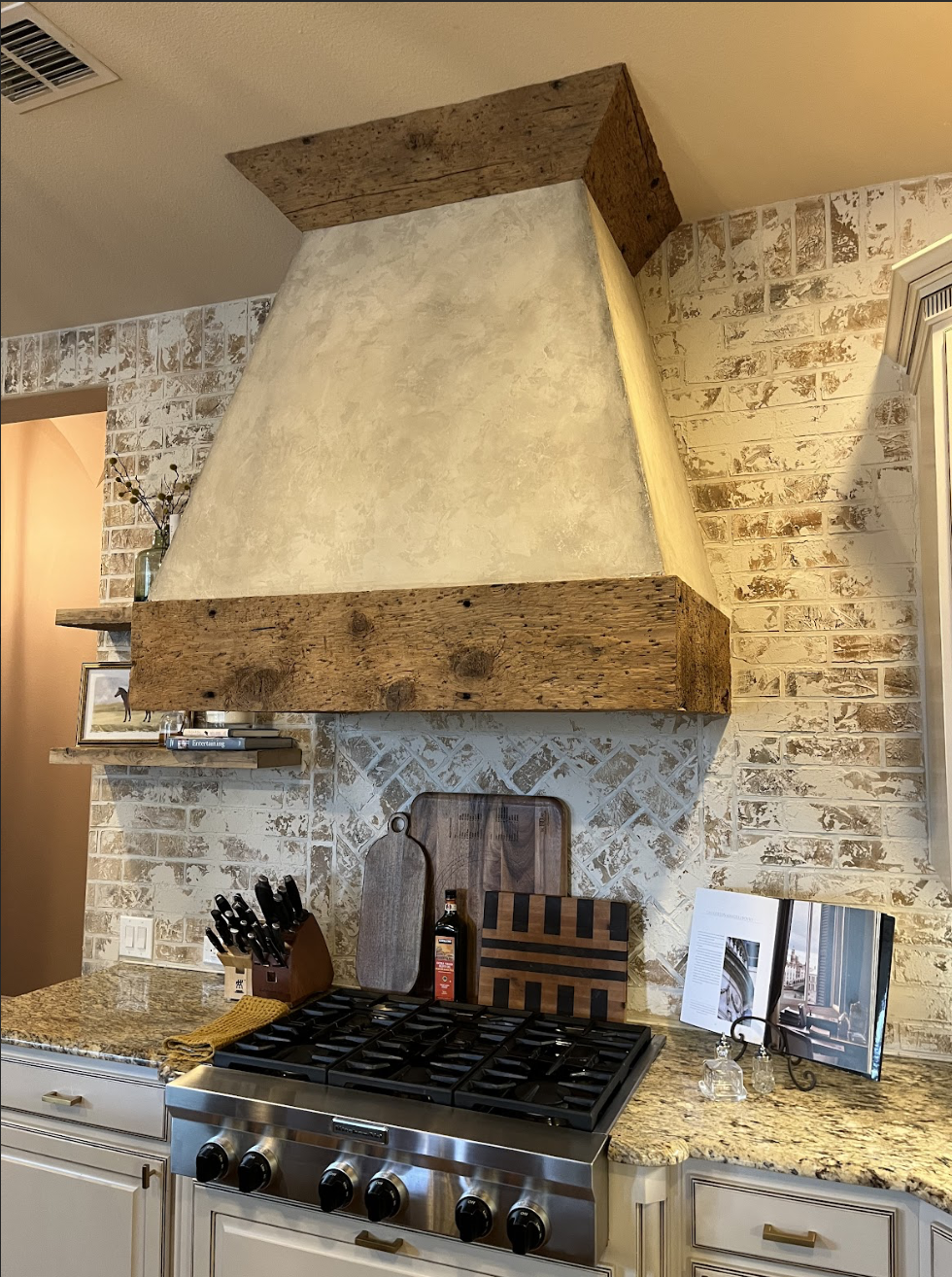 Kitchen with exposed brick walls, granite countertops, stainless steel stove, wooden hood vent, and decorative items including cutting boards, knives, and a cookbook stand.