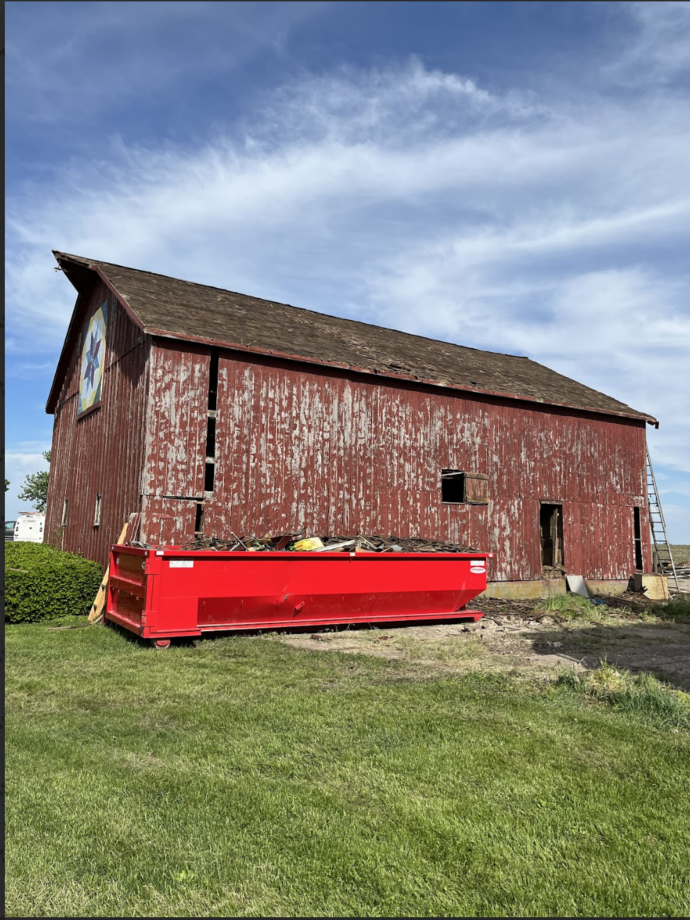 An old red barn with peeling paint and boarded-up windows, with a large red dumpster in front filled with debris, under a partly cloudy sky.