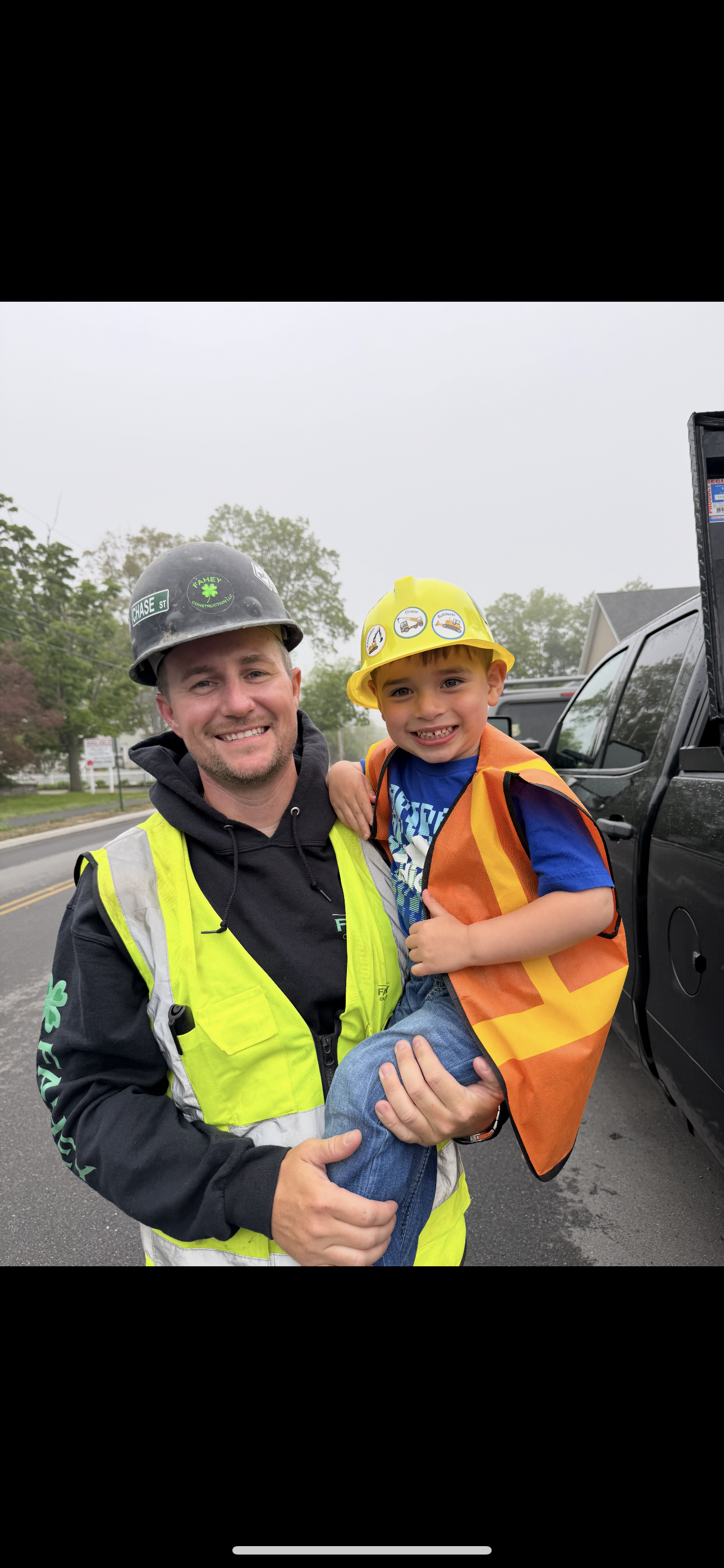 A man in a black helmet and high-visibility vest holding a smiling young boy in a yellow construction helmet and orange safety vest, standing outdoors on a street with cars and trees in the background.