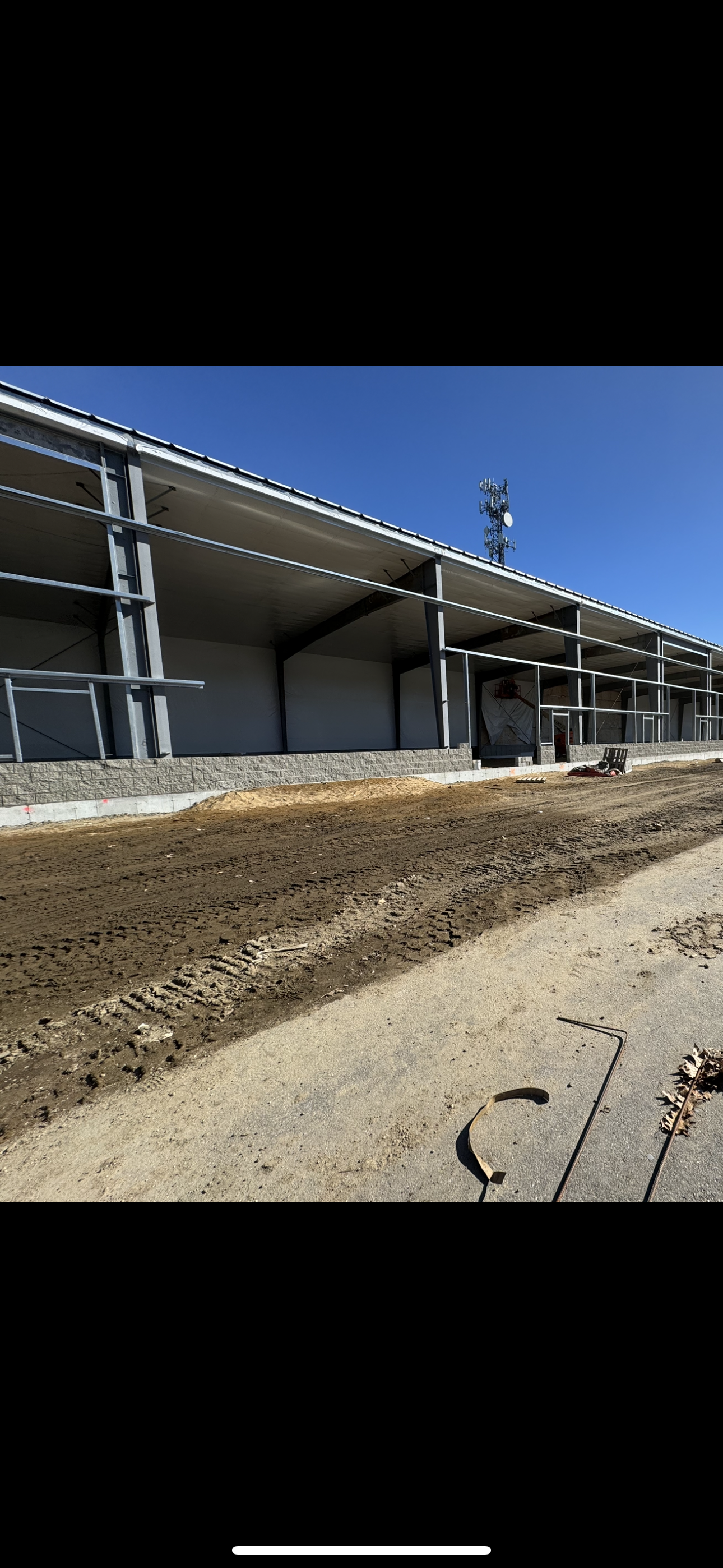 Construction site with a partially built building featuring exposed steel beams, a dirt ground with tire tracks, a clear blue sky, and construction tools on the ground.