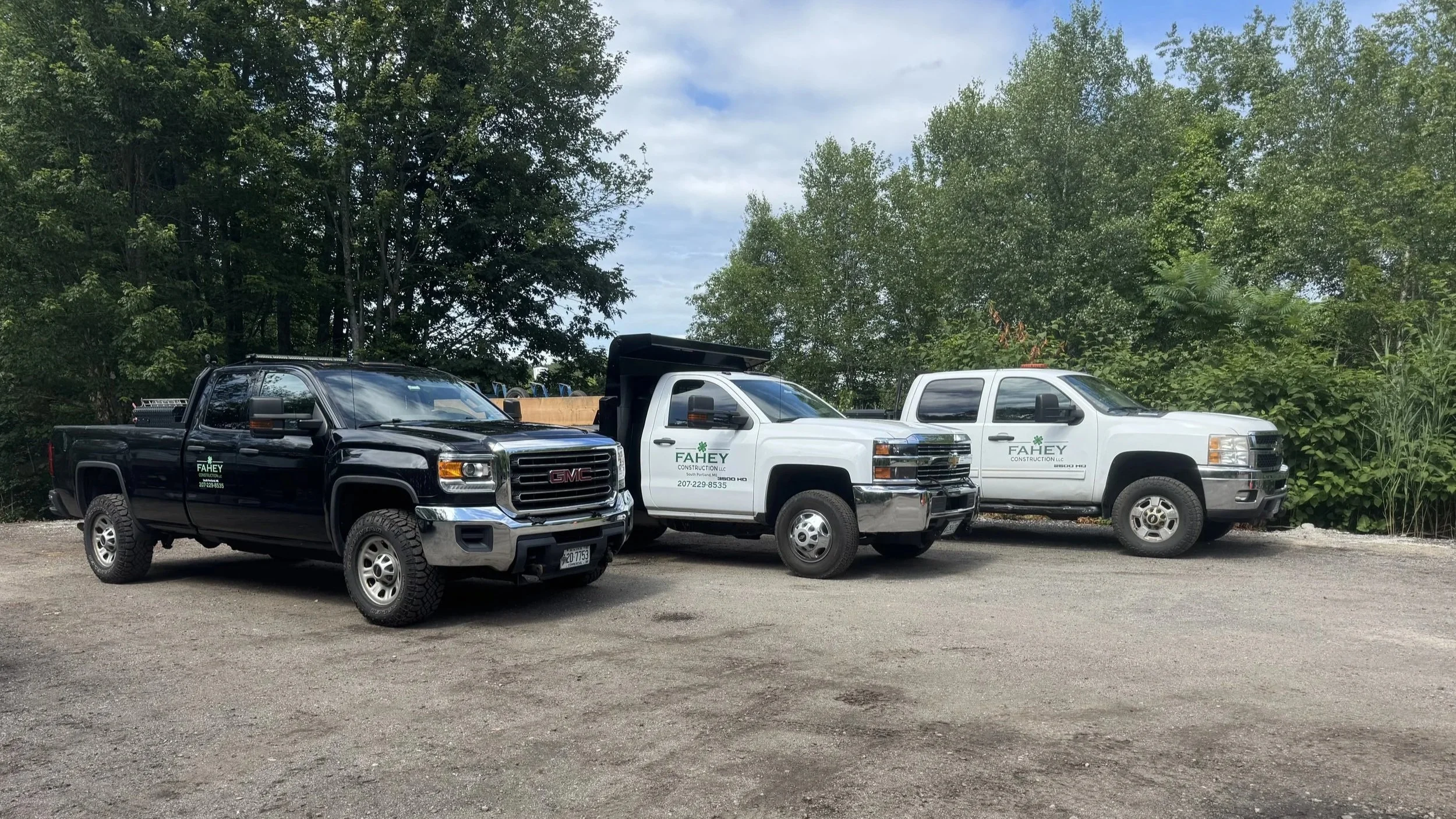 Three pickup trucks parked on a gravel lot with trees in the background. The trucks are black and two white, all with FAHEY Construction LLC logos on the doors.