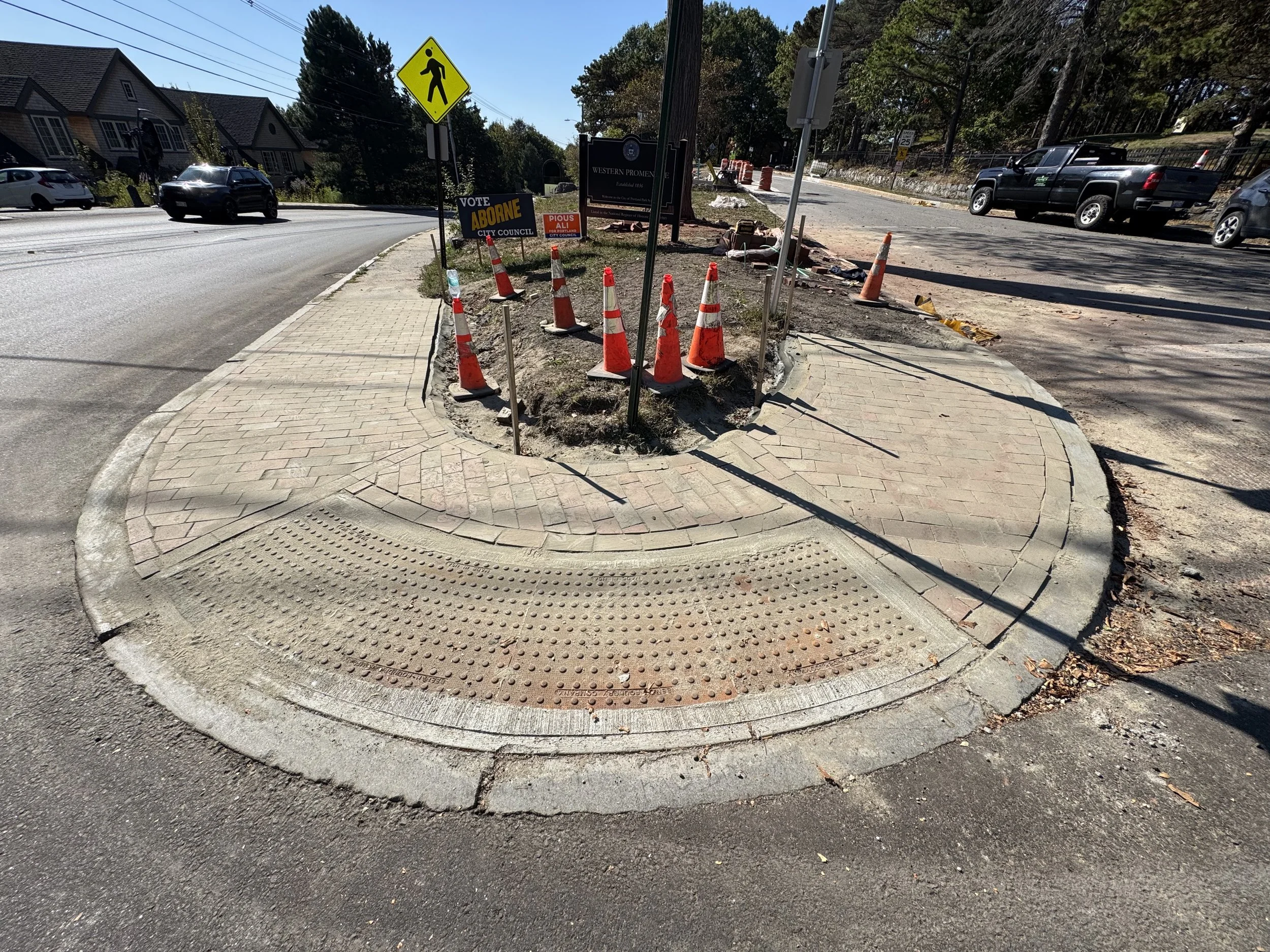 Construction area with orange cones and barriers on a sidewalk and street, with signs and parked cars in the background.
