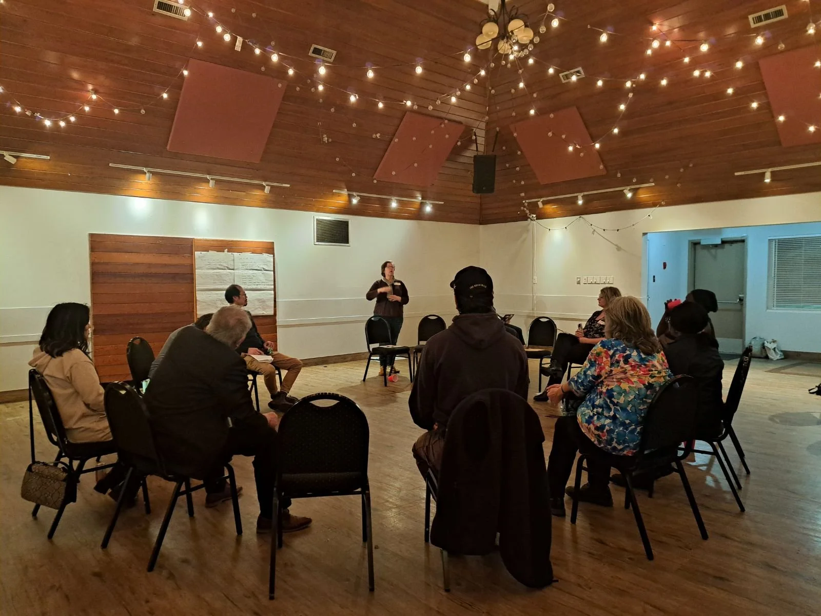 A small group of people is attending a presentation or meeting in a decorated wooden ceiling room with string lights. They are sitting on black chairs facing a woman standing and speaking at the front.