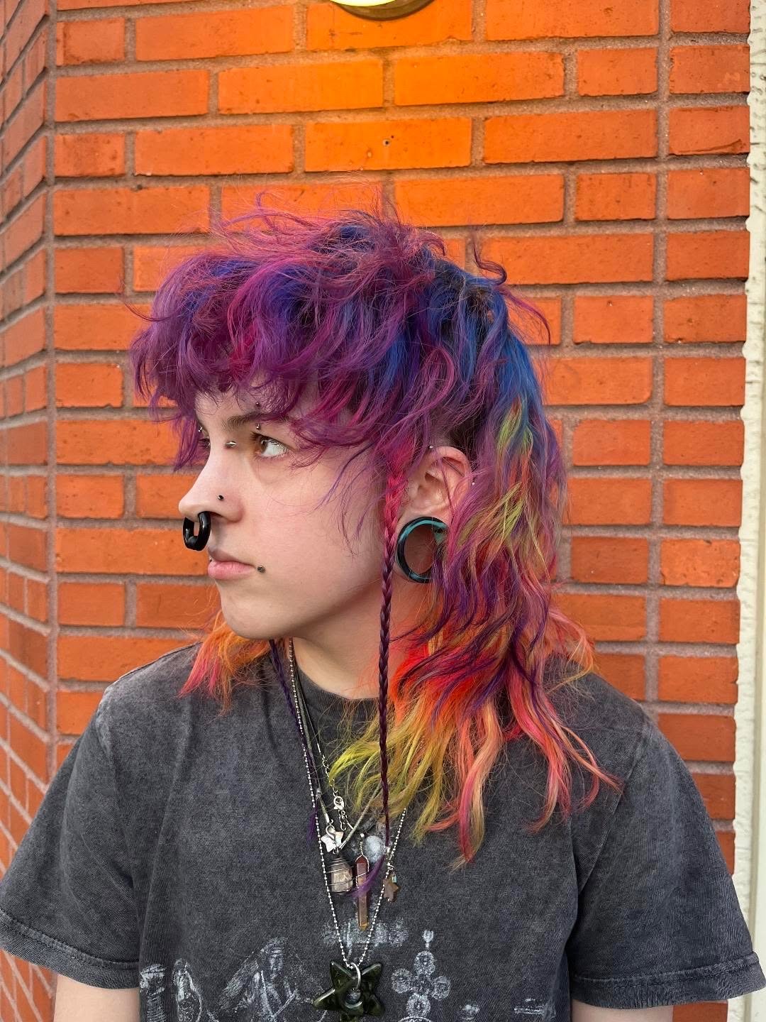Person with rainbow-colored, curly, multi-layered hair and several facial and ear piercings, standing against a brick wall.