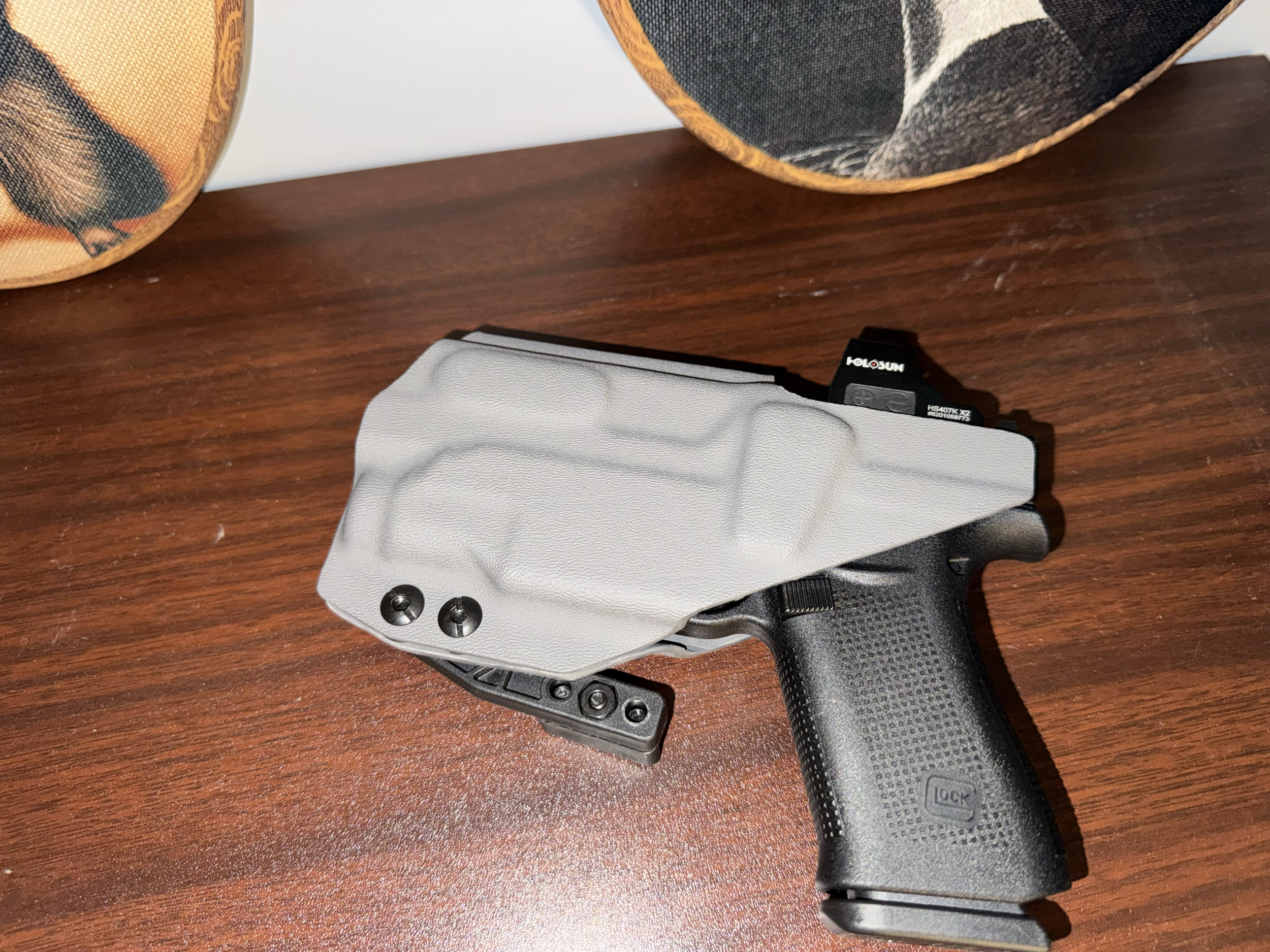 A handgun with a tan tactical holster on a wooden surface, with two decorative fabric items in the background.