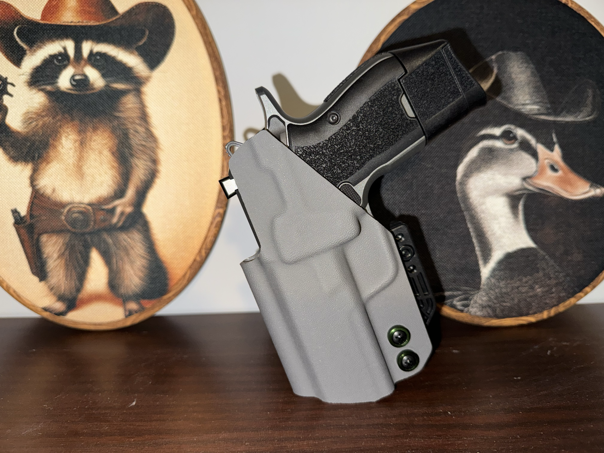 Close-up of a handgun with a textured grip resting on a wooden surface between two decorative round items with raccoon and fox images.