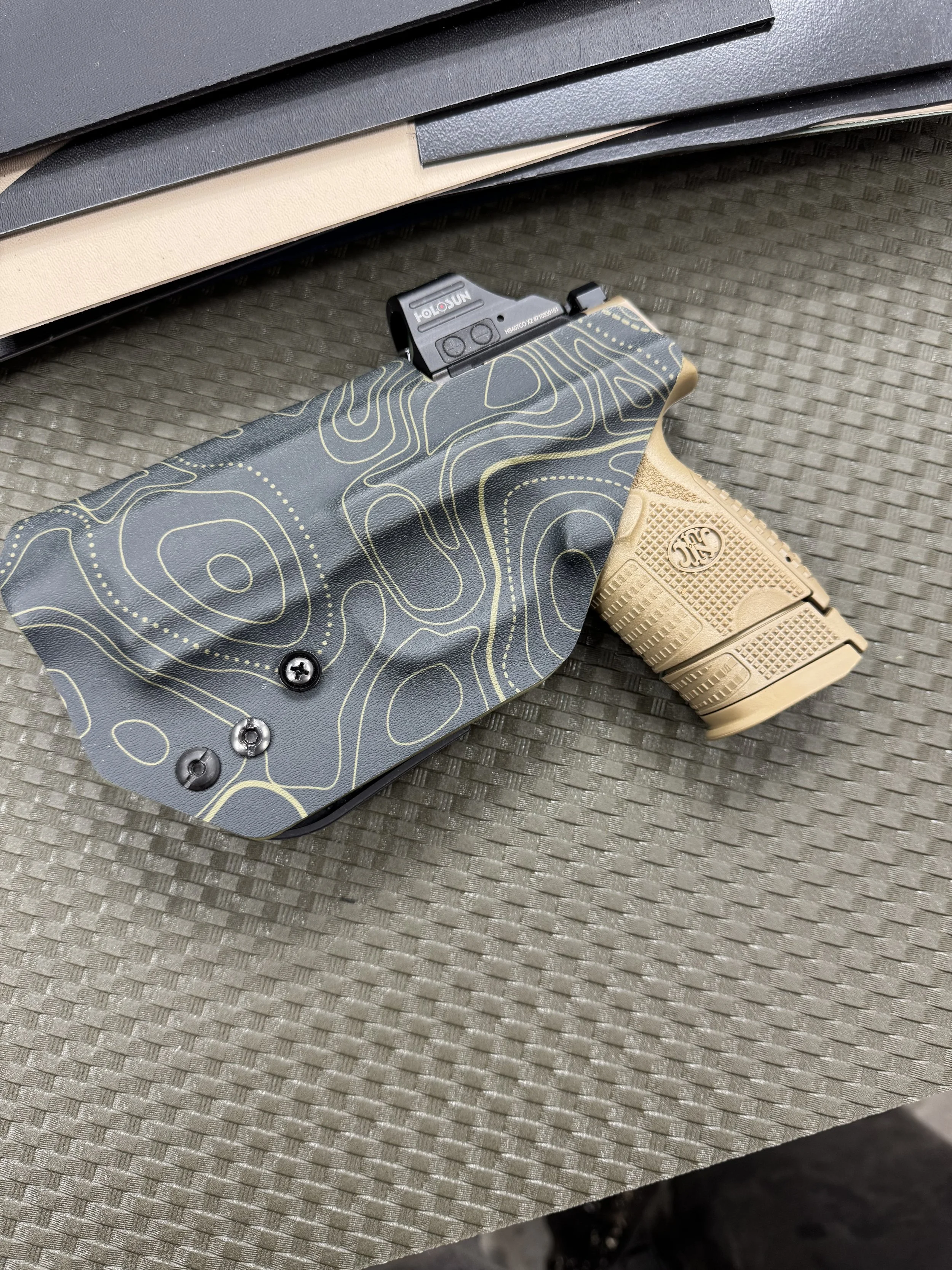 A pistol with a tan grip and a black slide resting on a textured surface. The slide has a mounted red dot sight, and the grip features a logo.