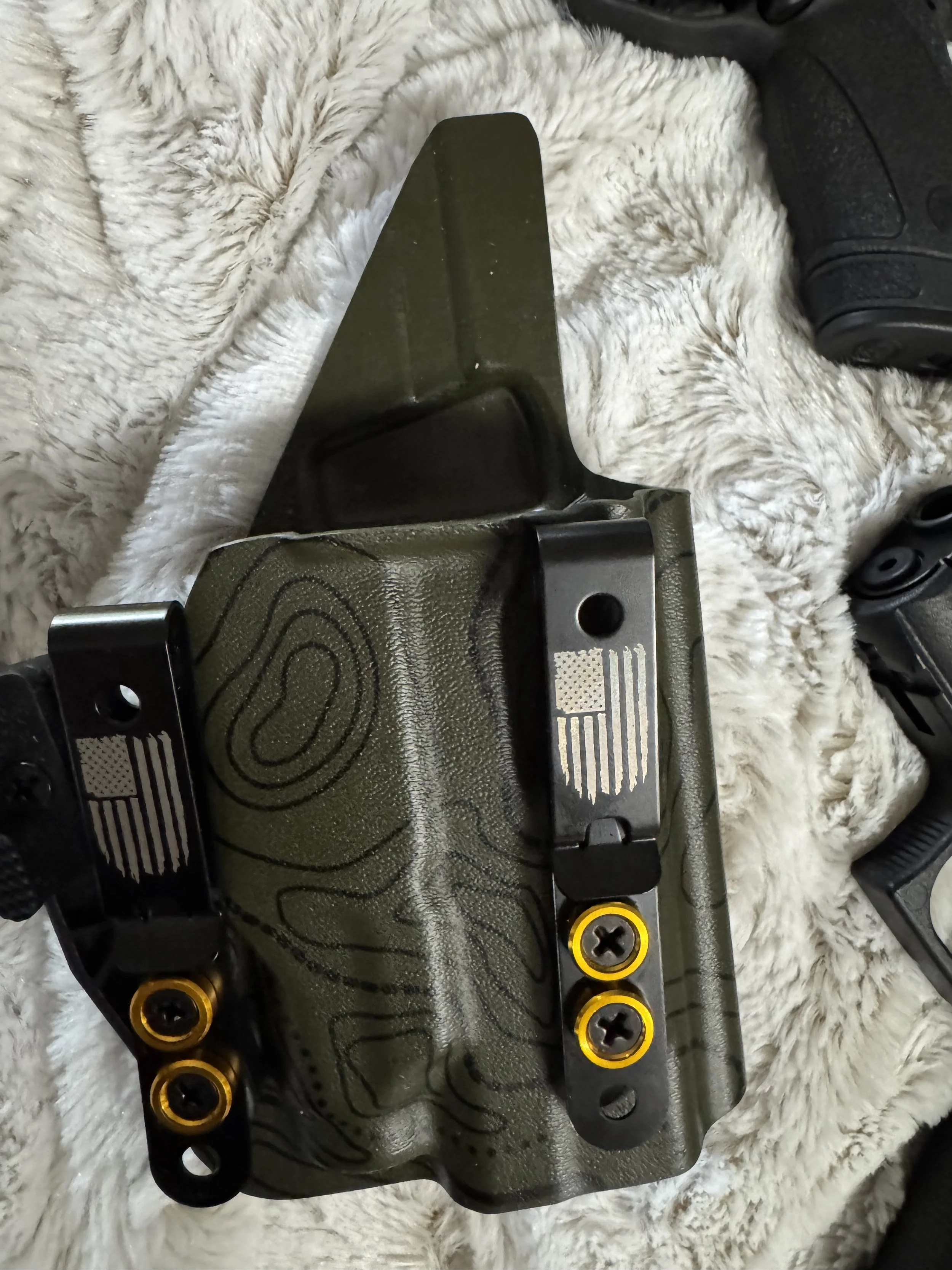 Close-up of a piece of military or tactical equipment, possibly a weapon attachment, with a textured black and green surface, gold accents, and screws, lying on a fluffy white surface.