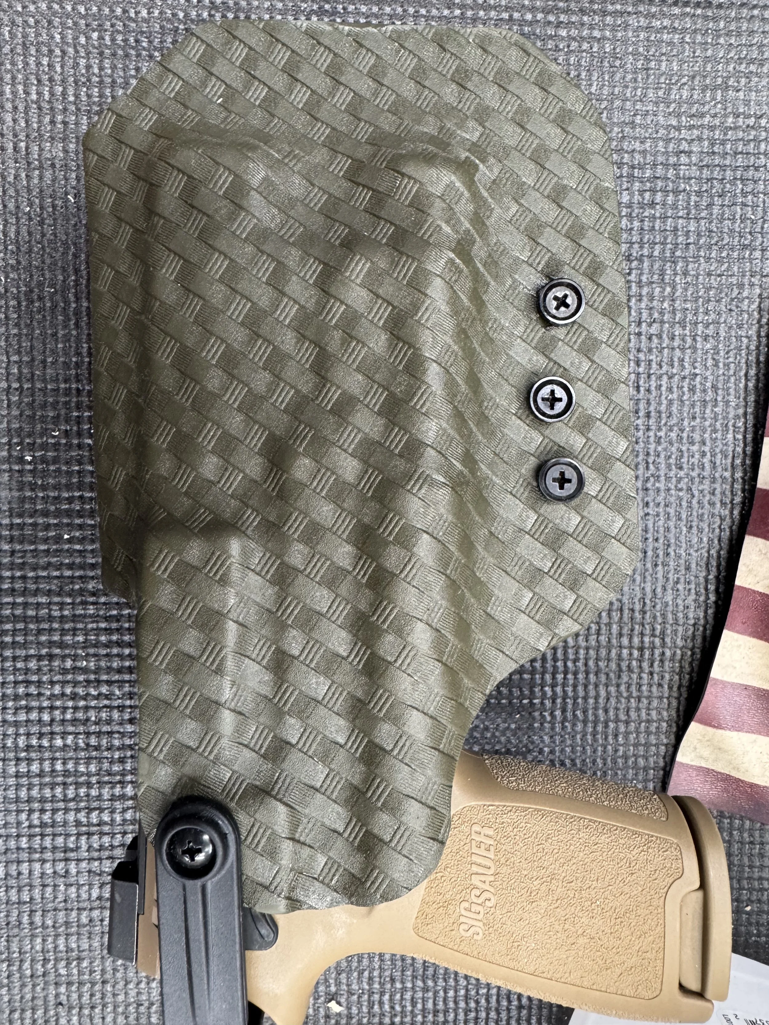 A close-up view of gun accessories including a textured rifle grip, a beige gun magazine with the brand name 'SHIELD' on it, and a black mounting plate with screws on a textured surface.