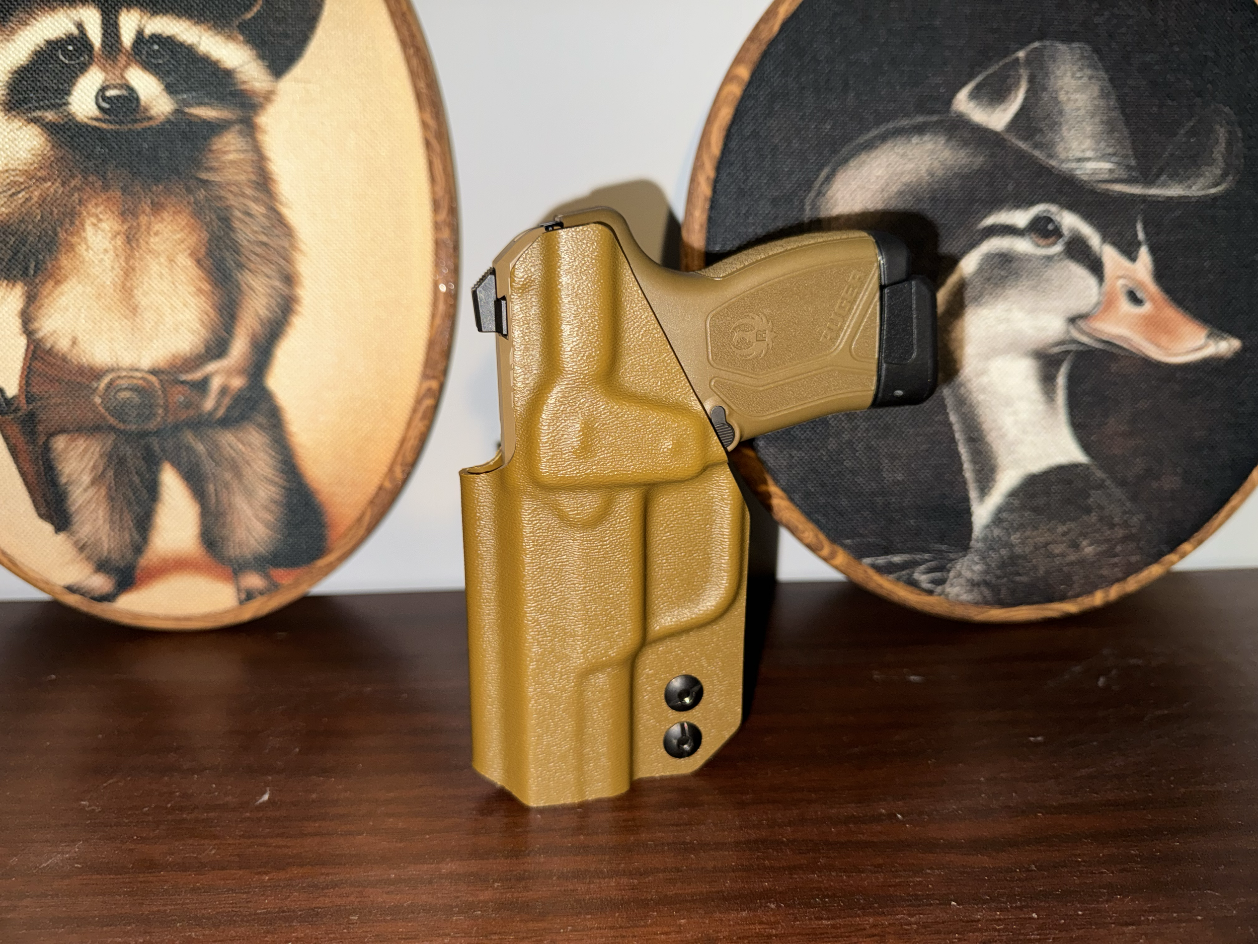 A tan and black handgun with a textured grip, positioned in front of two round wooden plaques with animal images, on a dark wooden surface.