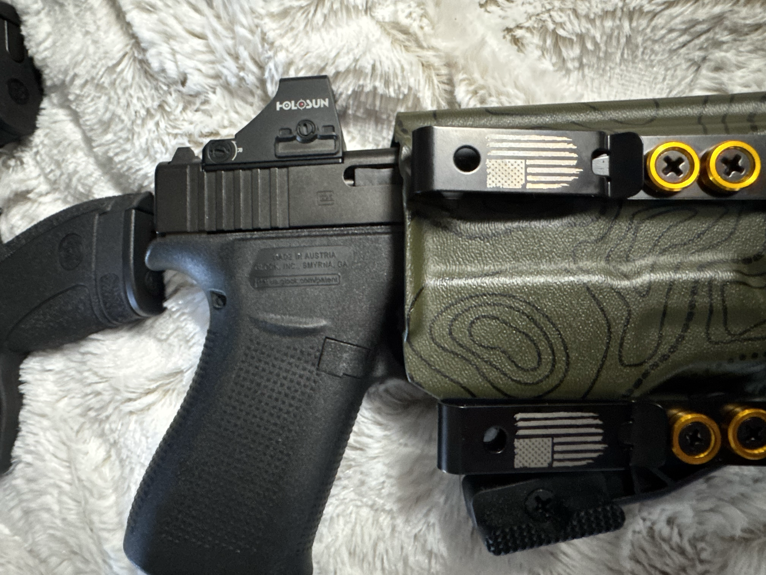 Close-up of a handgun's slide, with a holographic sight mounted on top, and part of another gun with a black grip and textured surface visible in the background. The guns are resting on a soft, white, fluffy surface.