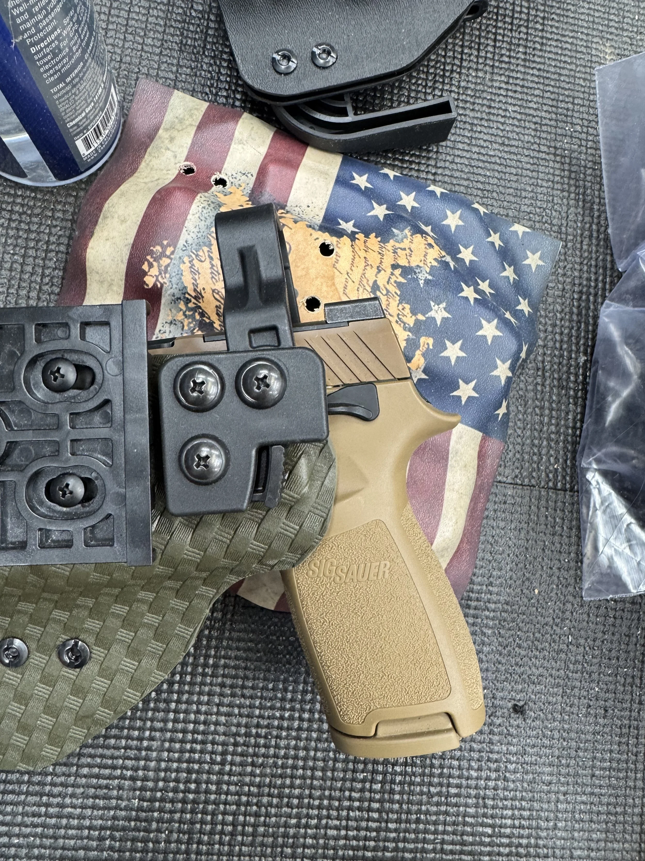 A handgun with a tan grip, a black attachment, and the SIG Sauer logo on the grip, placed on a textured surface with military-style camouflage mats, a American flag printed mat, and objects around.