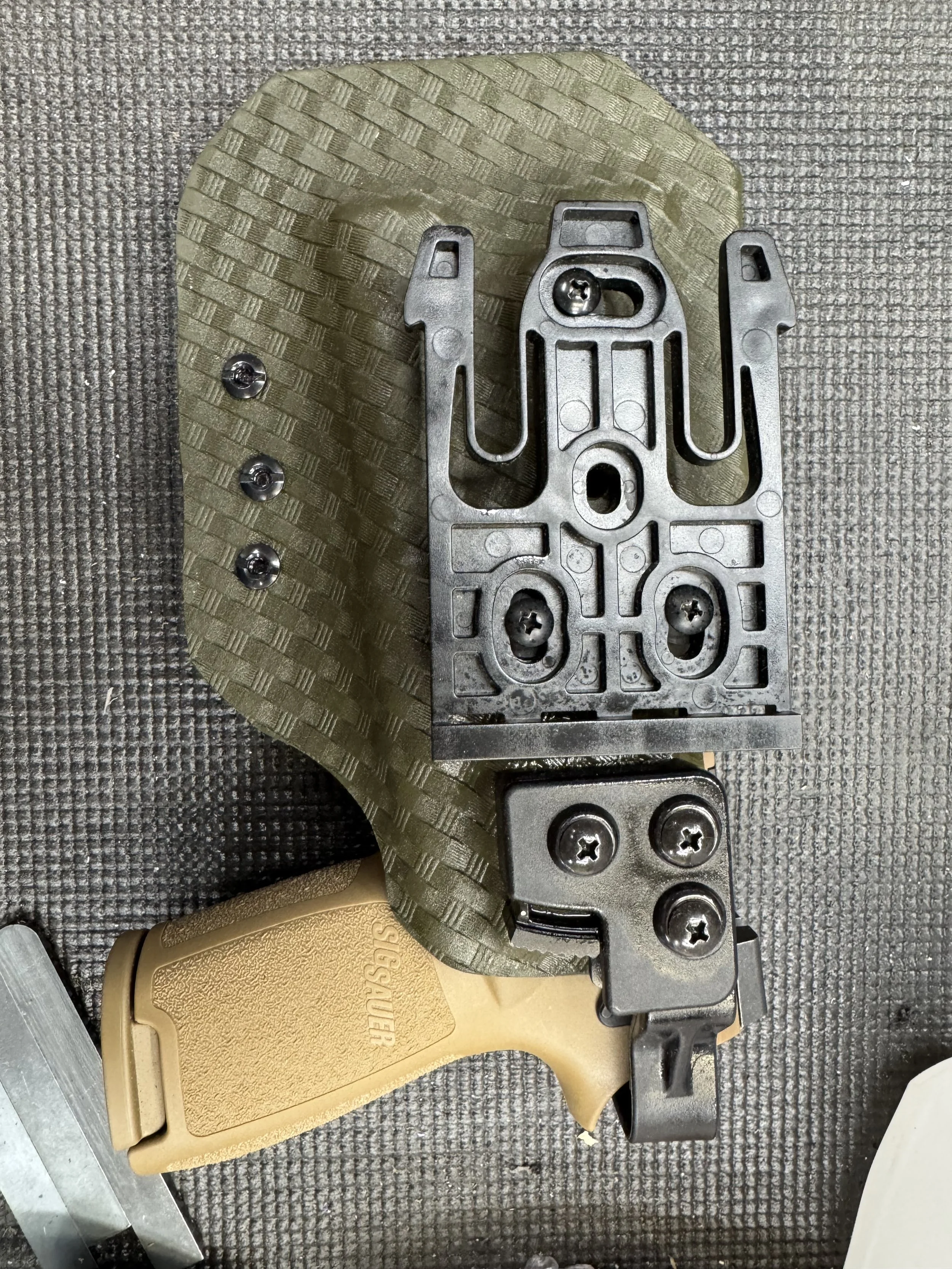 A handgun with a tan grip and black slide, mounted with a black frame, placed on a textured gray surface. The handgun is partially covered by a black metal mounting hardware and positioned next to a textured greenish cloth.
