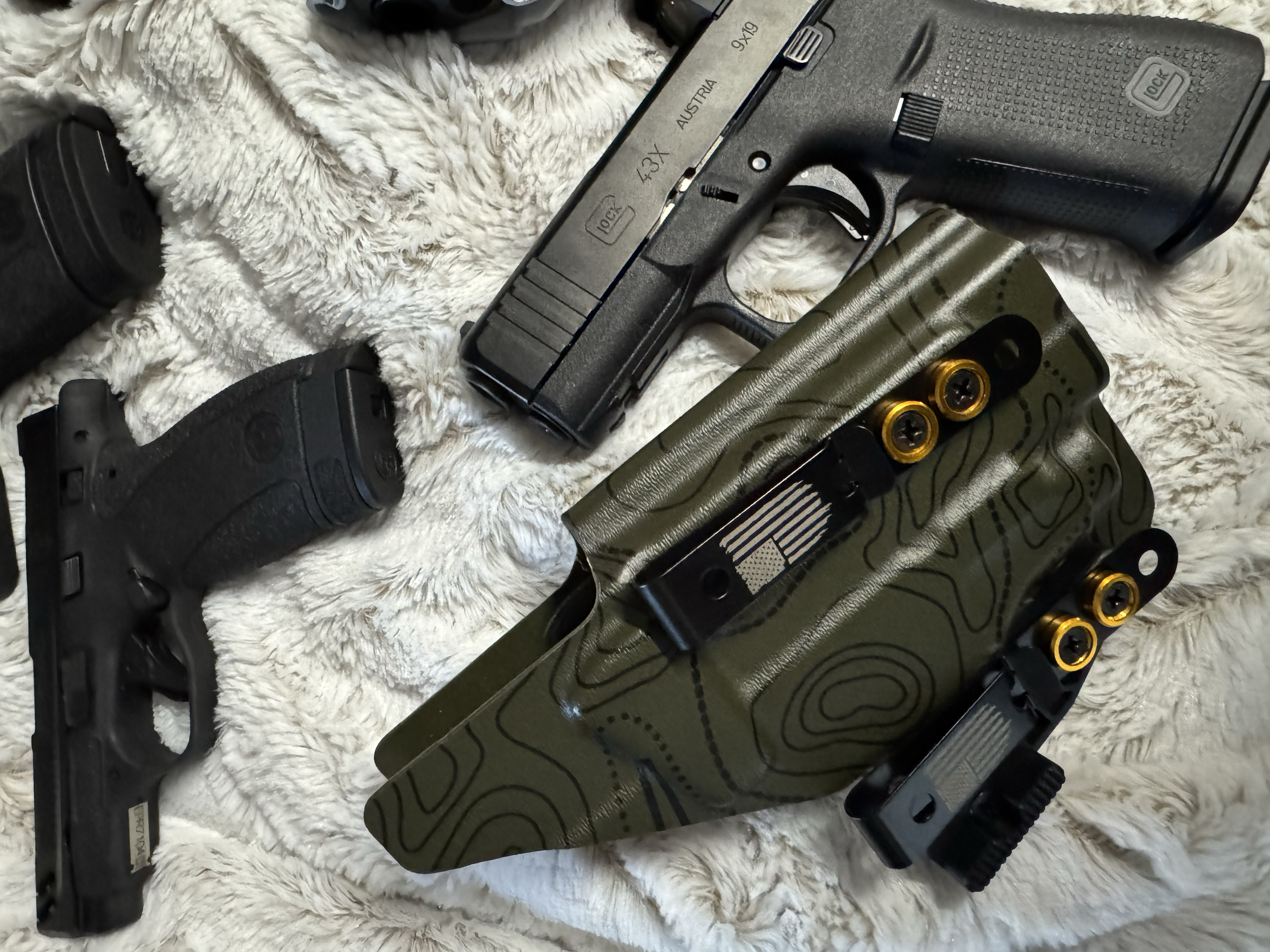 A semi-automatic handgun with a black slide and grip, a green tactical vest with yellow ammunition rounds, and black handgun holsters on a white fluffy surface.