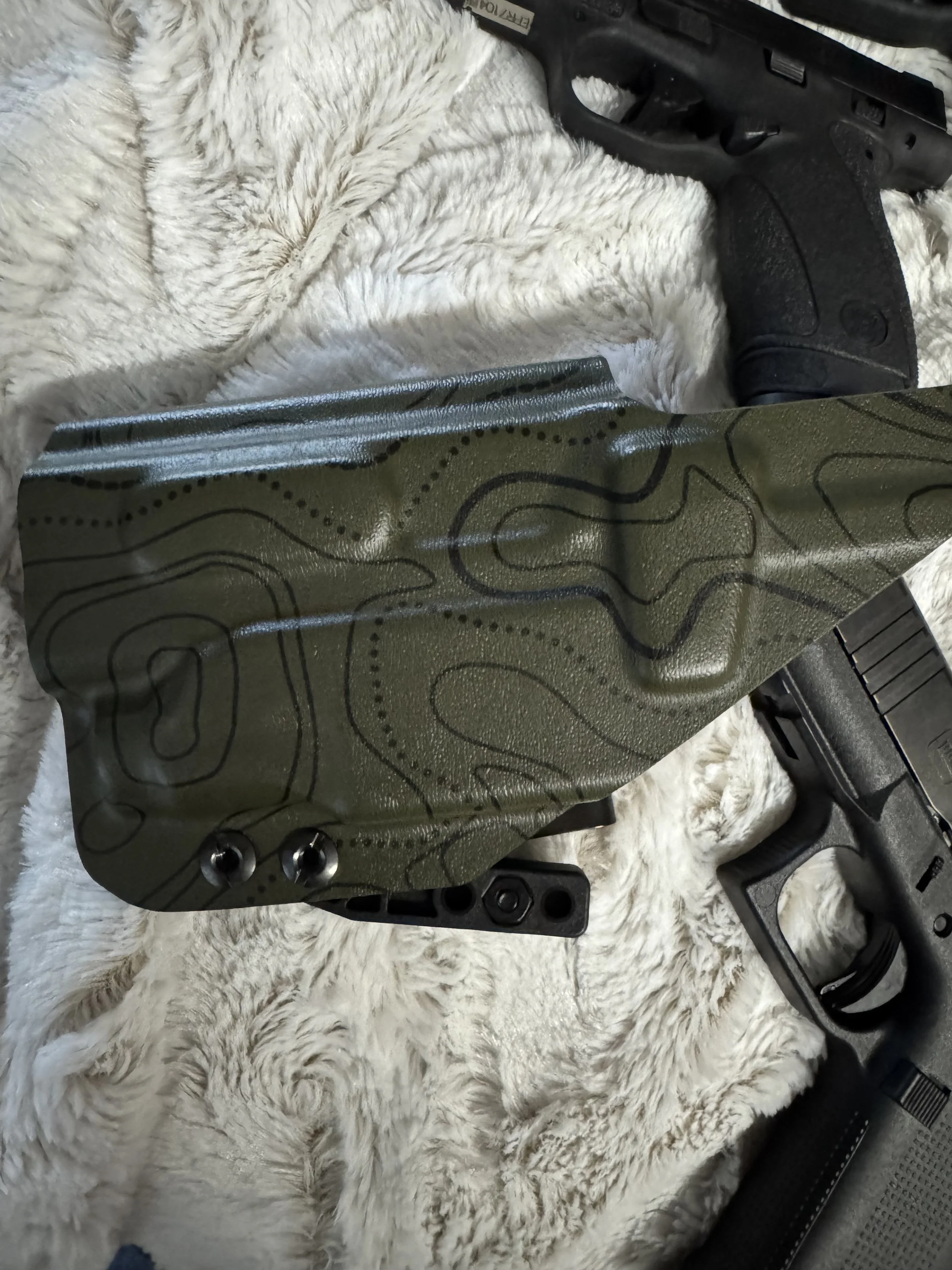 A black handgun and a green tactical knife with topographical markings are resting on a white, fluffy, fur-like surface.