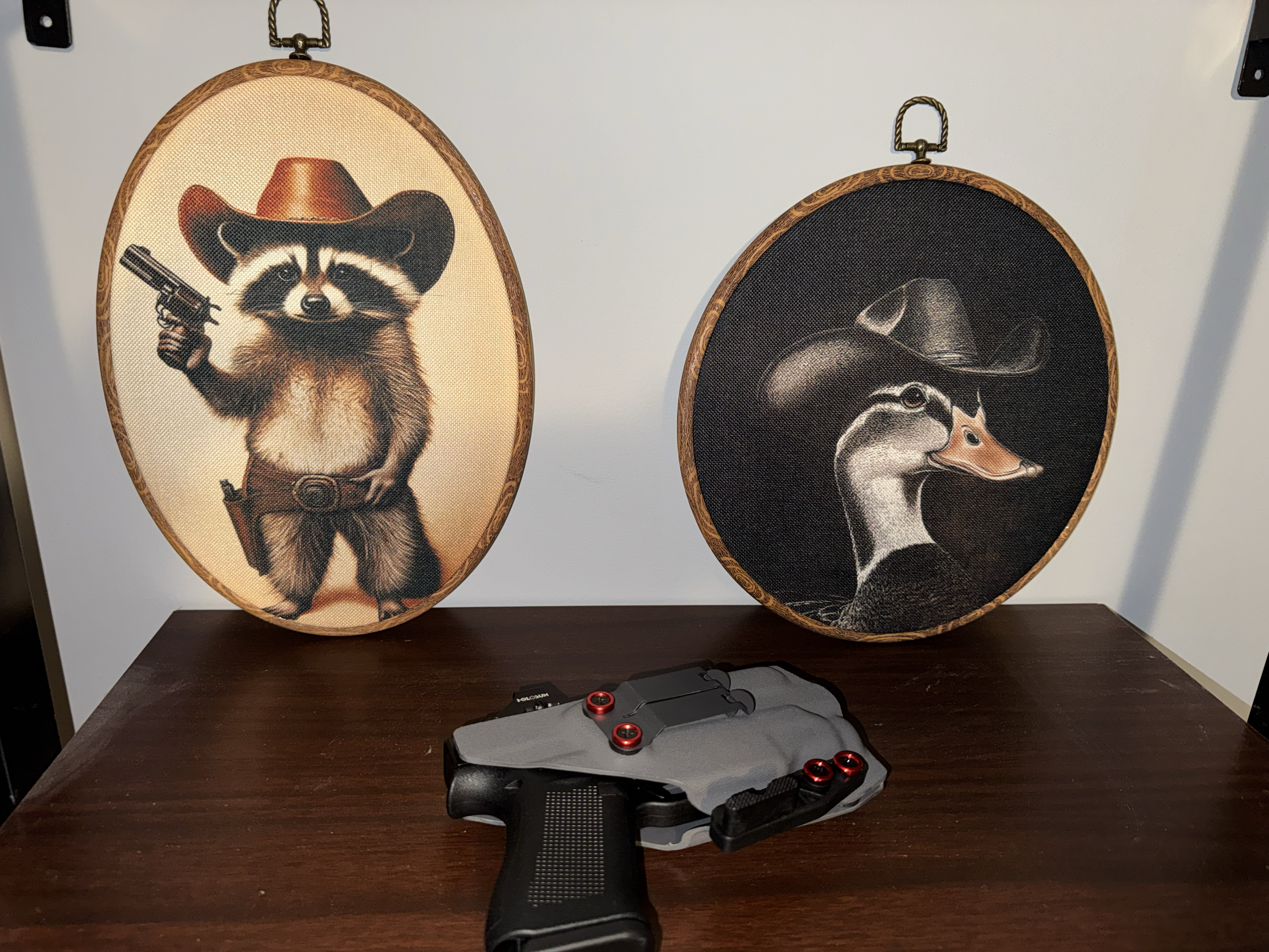 Two oval framed artistic prints of animals wearing western hats, one raccoon and one duck, mounted on a white wall. A black and gray handgun model is placed on a dark wooden table in front of the prints.