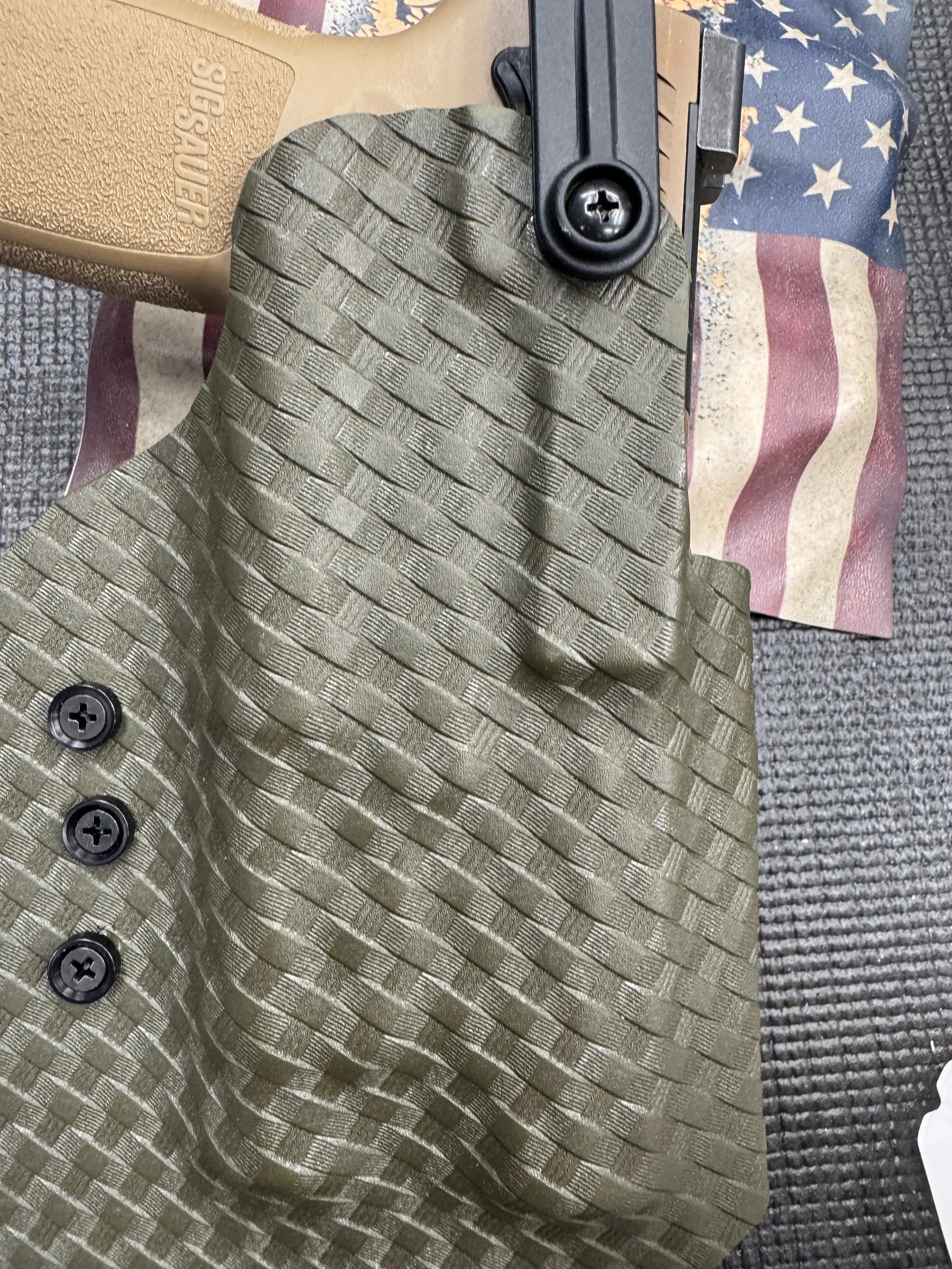 Close-up of tactical gear including a tan plastic holster with 'CRUSADER' embossed on it, a green textured glove, and part of a firearm with screws, placed on a partially visible American flag-themed mat.