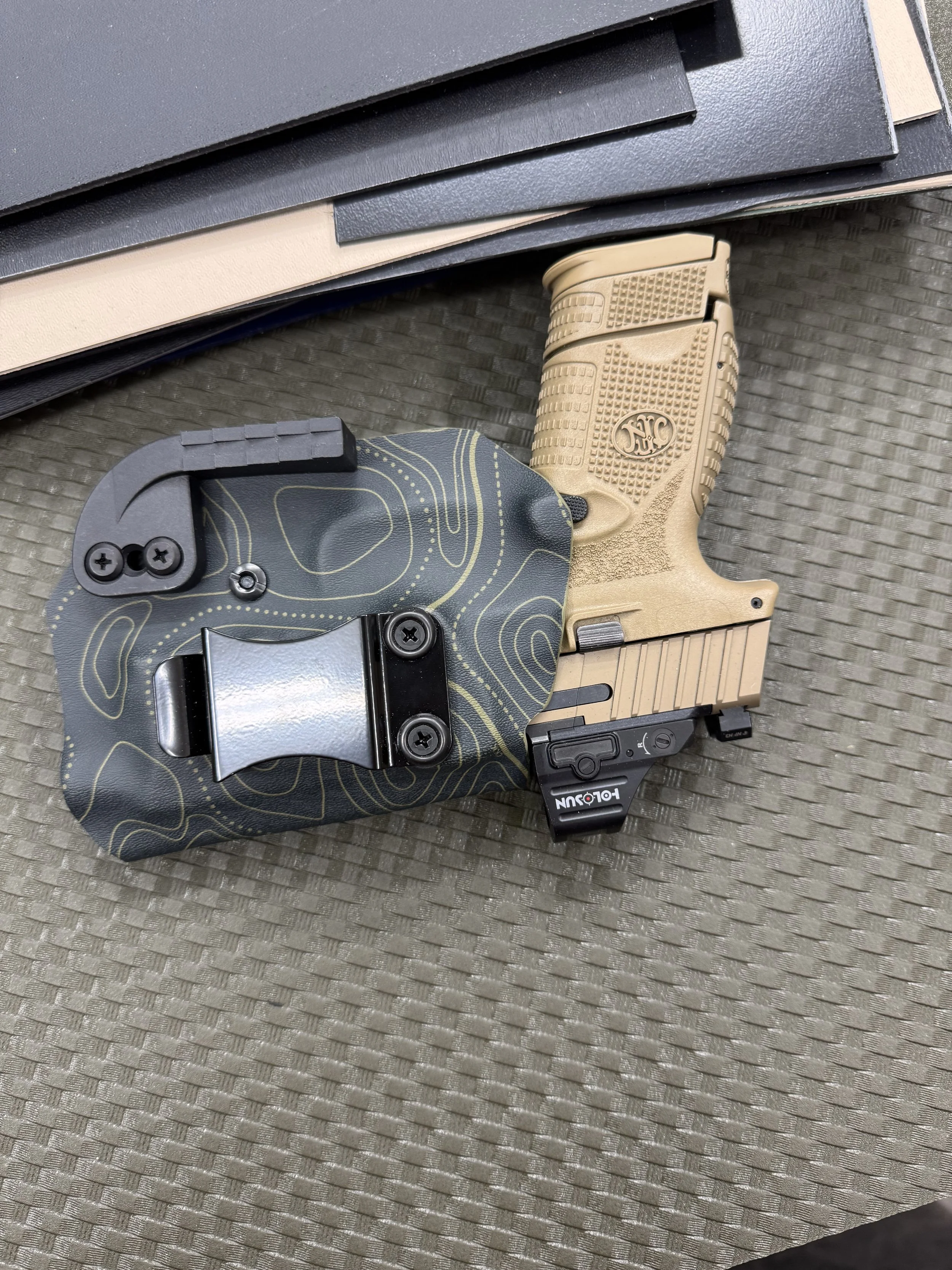 A tan Glock handgun with textured grip sitting on a gray surface, with a black and yellow patterned holster and several black and gray rectangular objects overlapping each other.